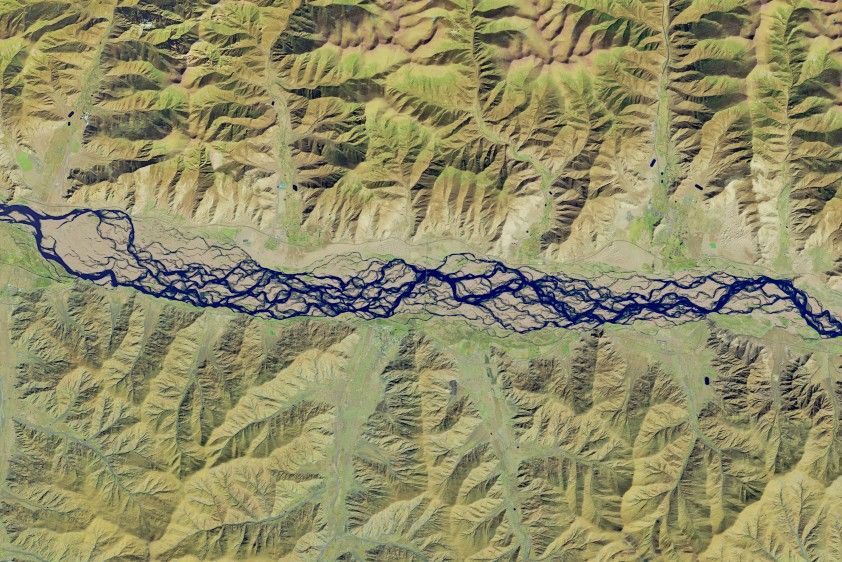 A false-color satellite image shows numerous intertwining channels of blue water spanning the middle of the scene from left to right. The river is bordered on the top and bottom by mountainous terrain, which appears tan and light green.