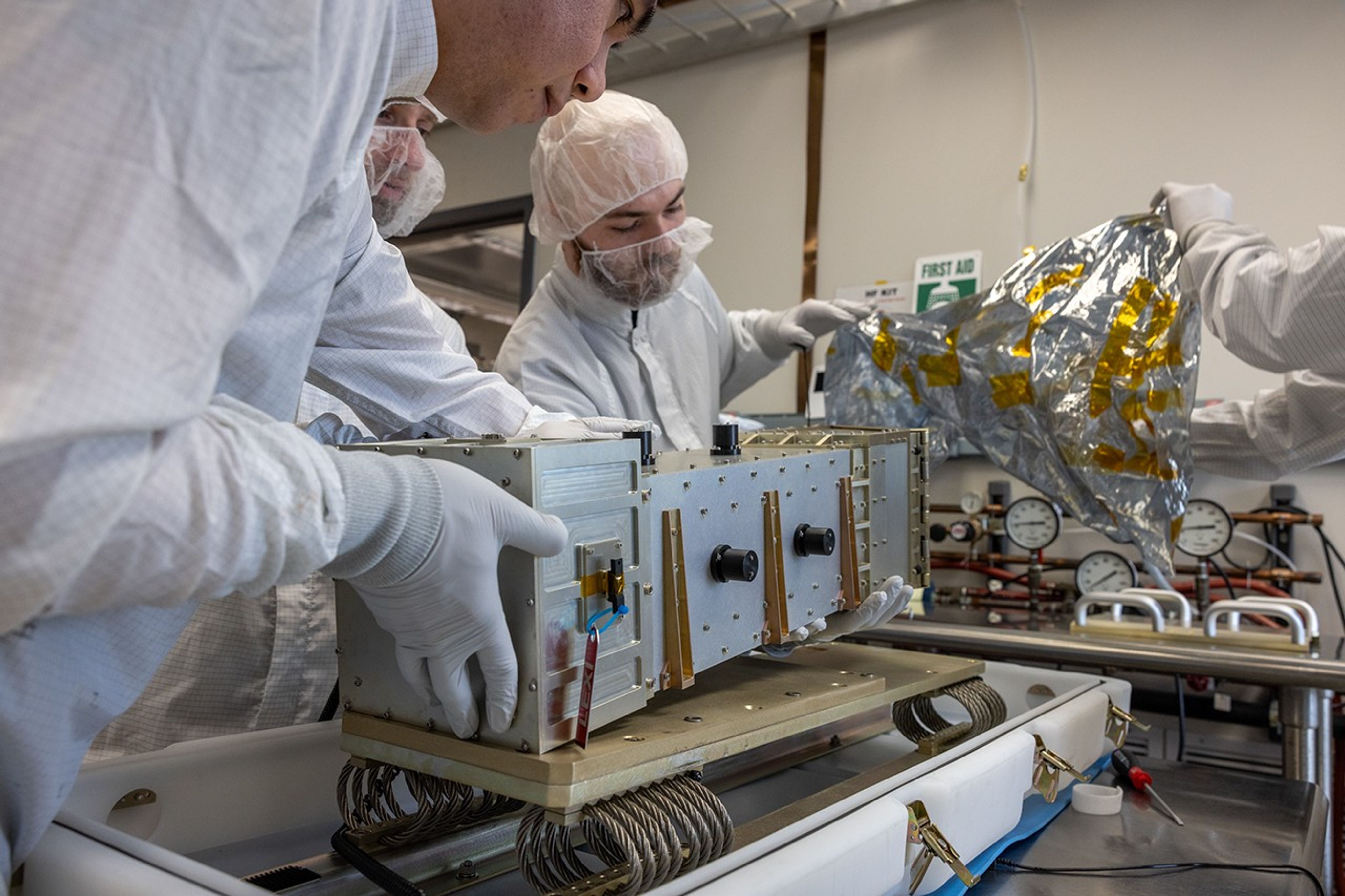 Several people dressed in white protective clothing handle the LEXI X-ray instrument and a sheet of silvery metallic film. The instrument is a boxy, rectangular shape and mostly gray metal.