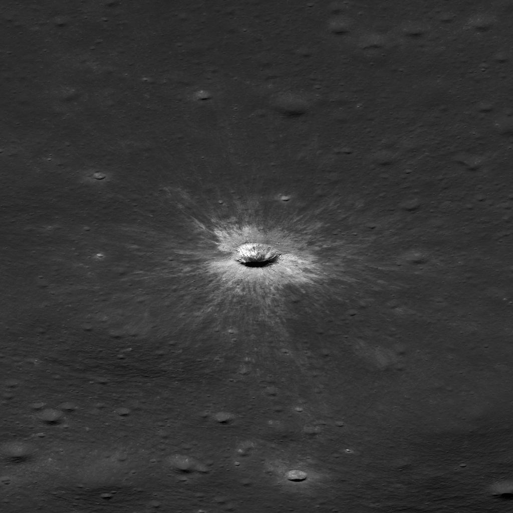Square black-and-white image of a dark, gently uneven surface scattered with small circular depressions. Near the center is a small oval crater with a bright raised rim and dark interior. Fine, bright streaks radiate outward from the crater across the surrounding surface.