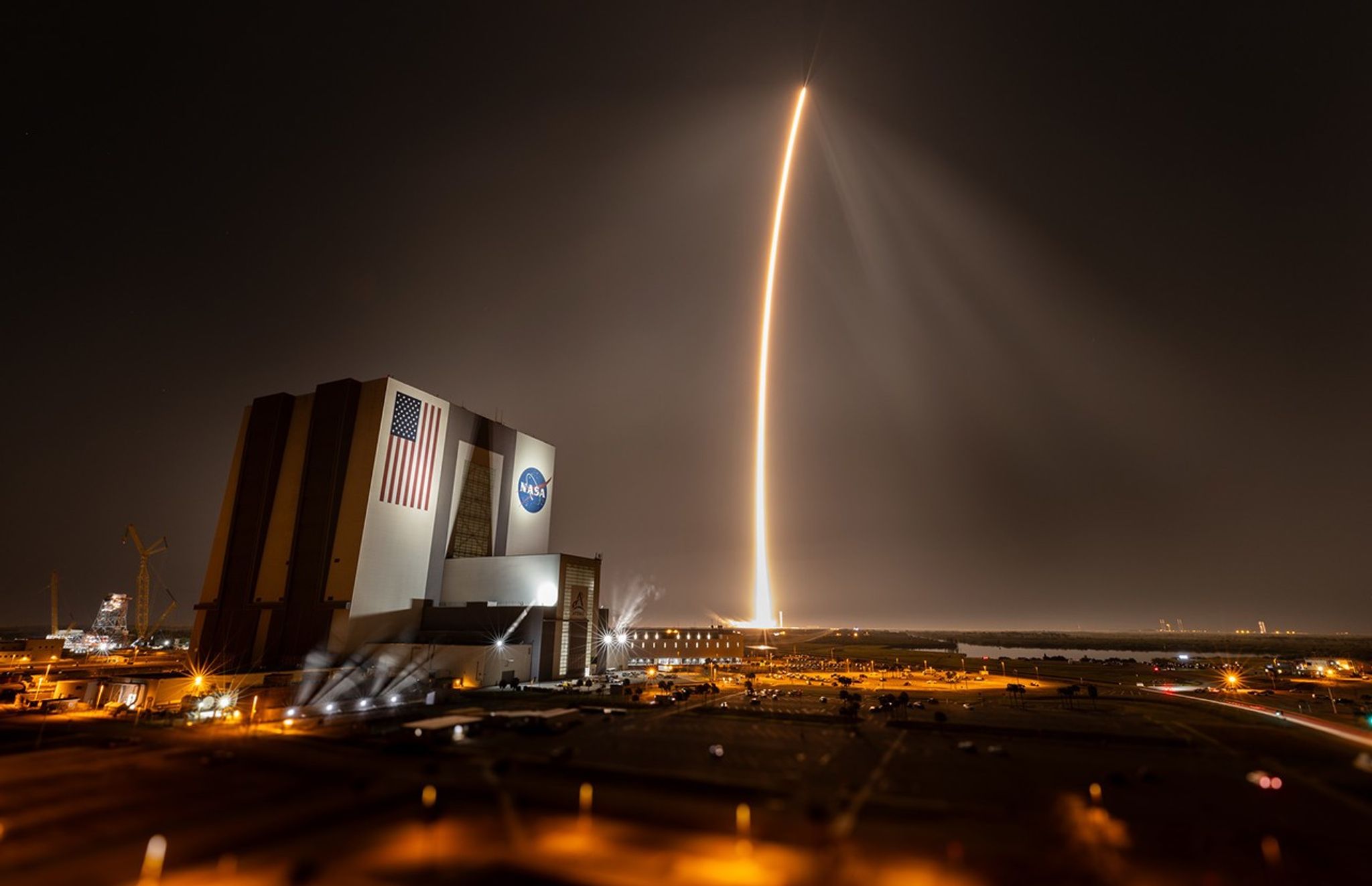 A bright reddish streak arcs over Kennedy Space Center. The Vehicle Assembly Building is to the left of the streak. It has a giant U.S. flag and NASA logo on the building.