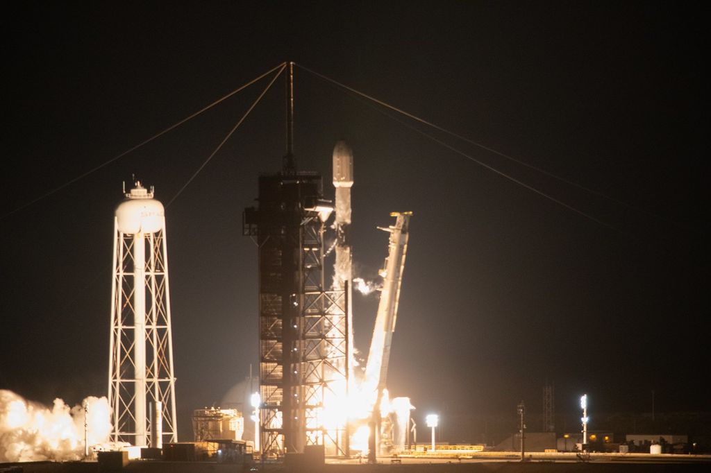 A white rocket with a white payload fairing on top lifts off the launch pad. White fire and smoke can be seen around the bottom of the rocket. A white water tower is off to the left.