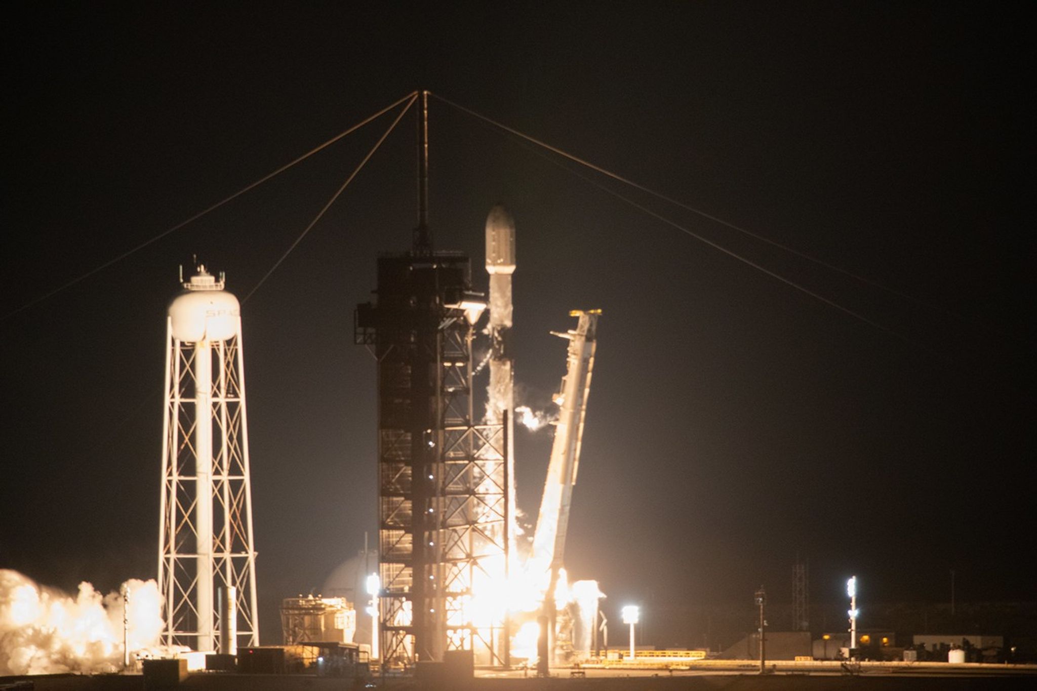 A white rocket with a white payload fairing on top lifts off the launch pad. White fire and smoke can be seen around the bottom of the rocket. A white water tower is off to the left.