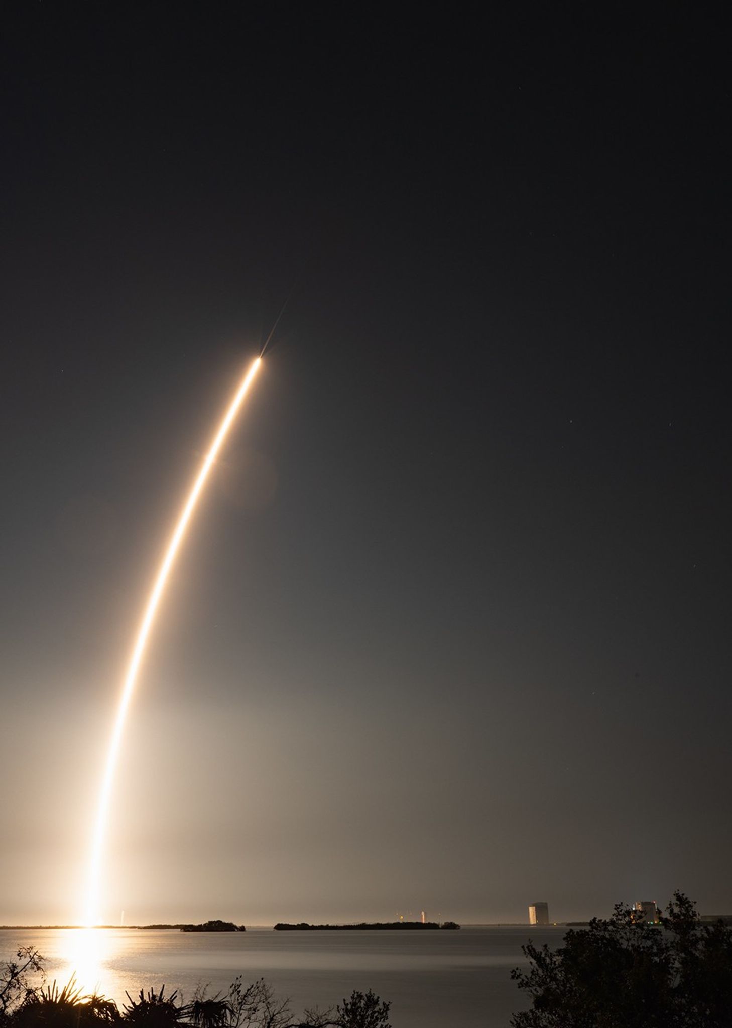 A reddish white streak arcs over Cape Canaveral leaving a bright reflection in the water below. A rocket is barely visible at the top of the streak.