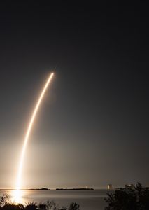 A reddish white streak arcs over Cape Canaveral leaving a bright reflection in the water below. A rocket is barely visible at the top of the streak.