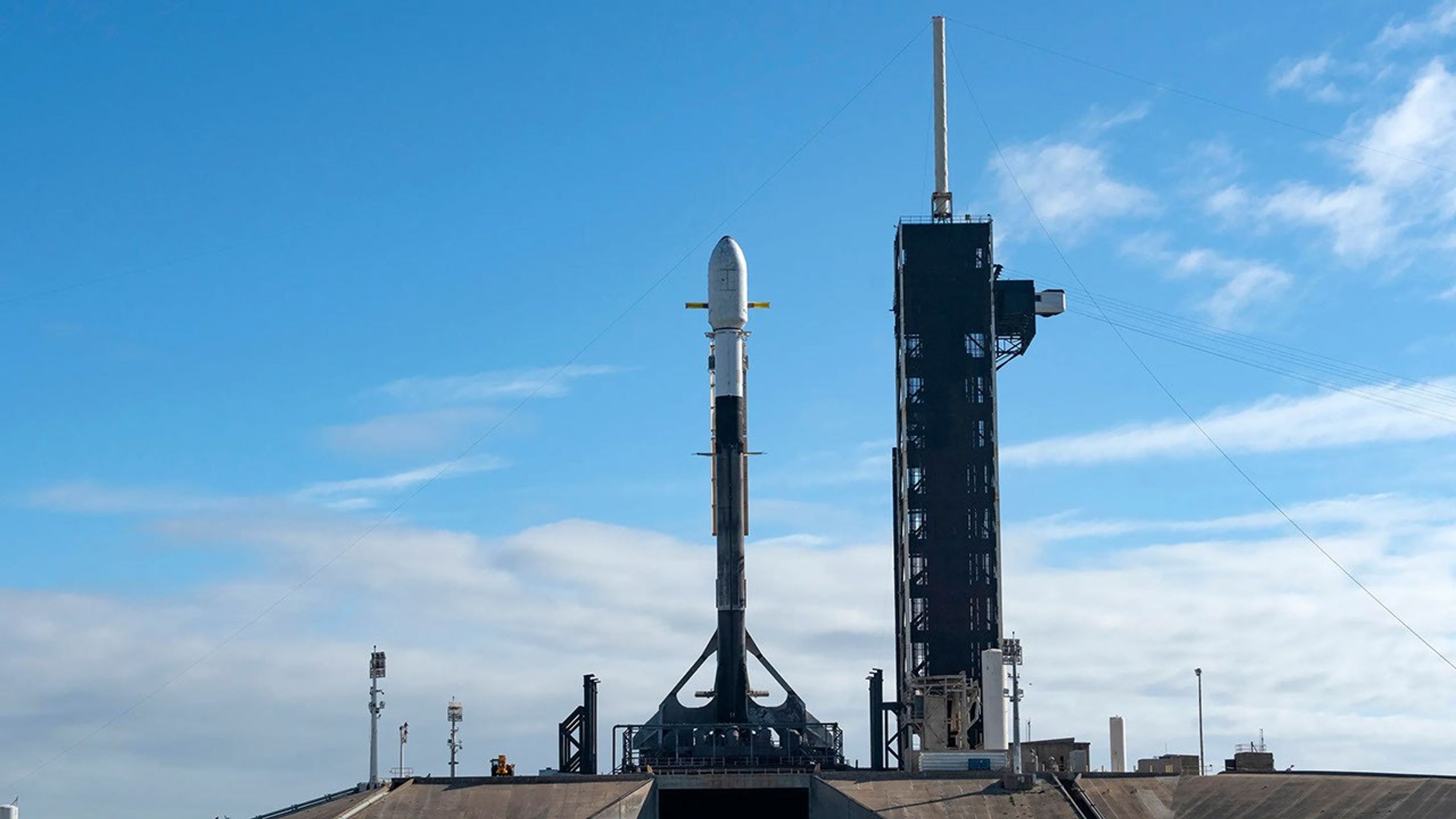 A rocket with a white capsule on top sits on a hill at the launch pad. The sky is blue with a few white, thick clouds near the horizone.