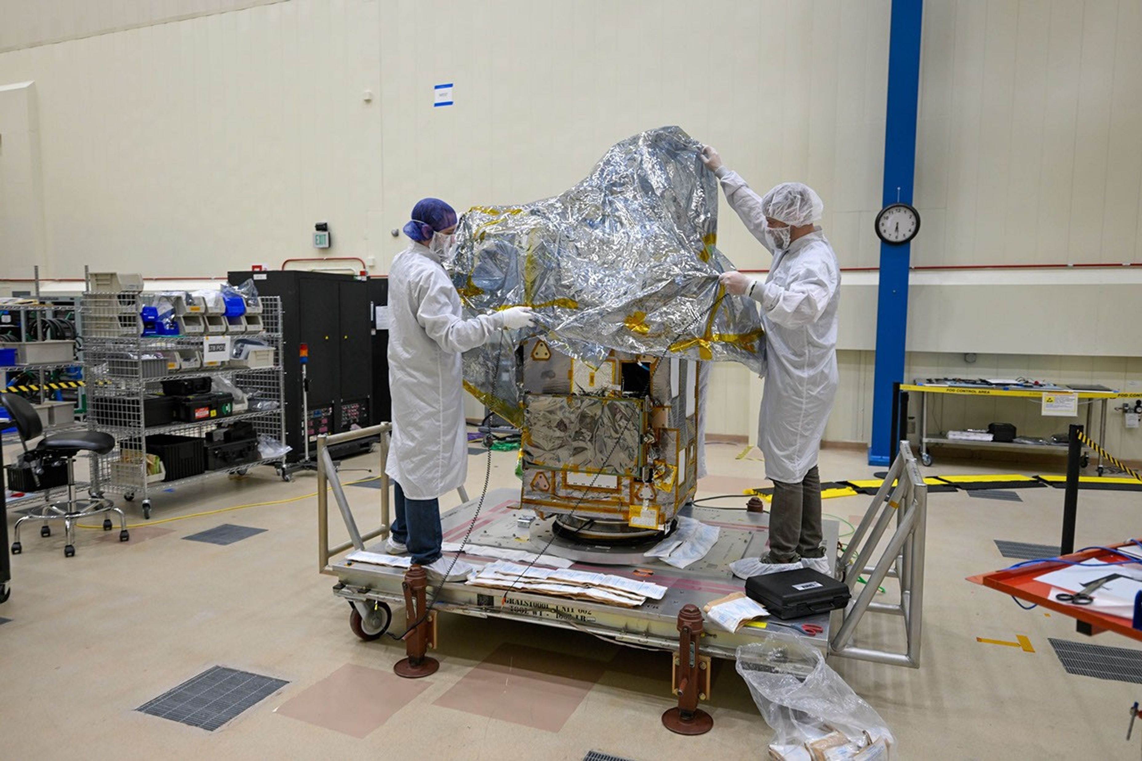 Two workers in protective clothing drape a cover over a spacecraft that is resting on a raised platform.