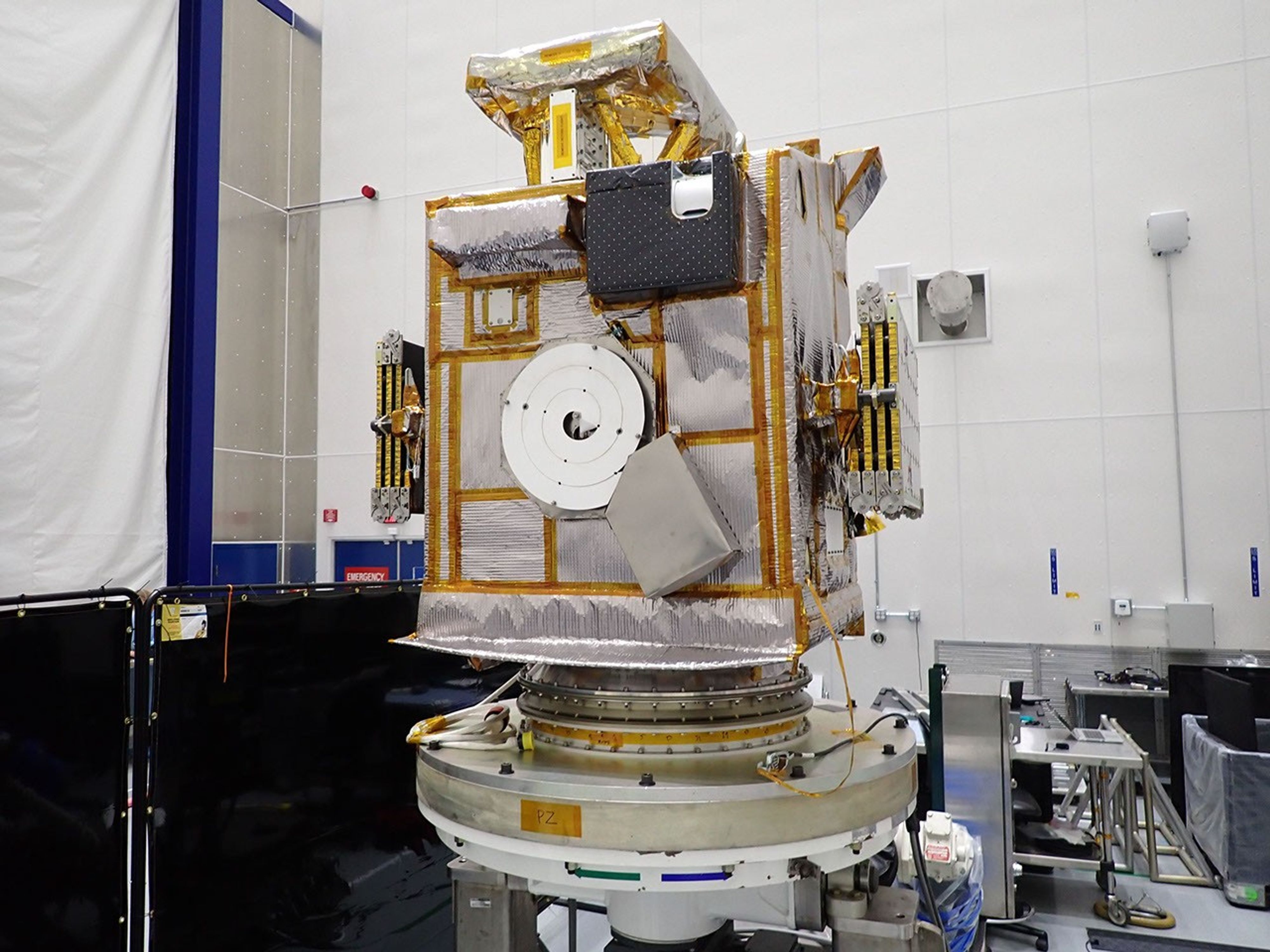 A box-shaped silver spacecraft sits on a servicing stand.