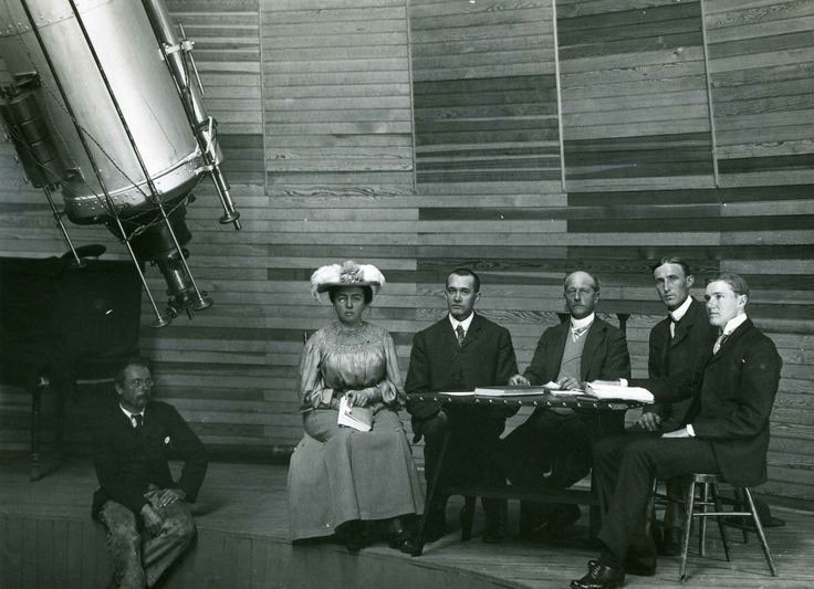 A vintage black and white photograph shows six people seated inside an observatory dome, with what appears to be horizontal wood-slat paneling on the wall behind them. A large, silvery-gray cylinder — the bottom end of the observatory telescope — dominates the upper-left corner of the image, extending from beyond the frame to the left-middle of the image, with a smaller eyepiece and controls protruding from the bottom. Below that, a man sitting on a ledge is dressed in a suit and tie, and looking off to the right. The other five are sitting in chairs on the ledge, to the right of him. From left to right, a woman wearing a long dress, a shiny blouse and a flowered hat is holding a book in her lap, and four men dressed in suits and ties are seated at a table. The man on the far right is looking off-image, to the left; the middle four people are looking at the camera.