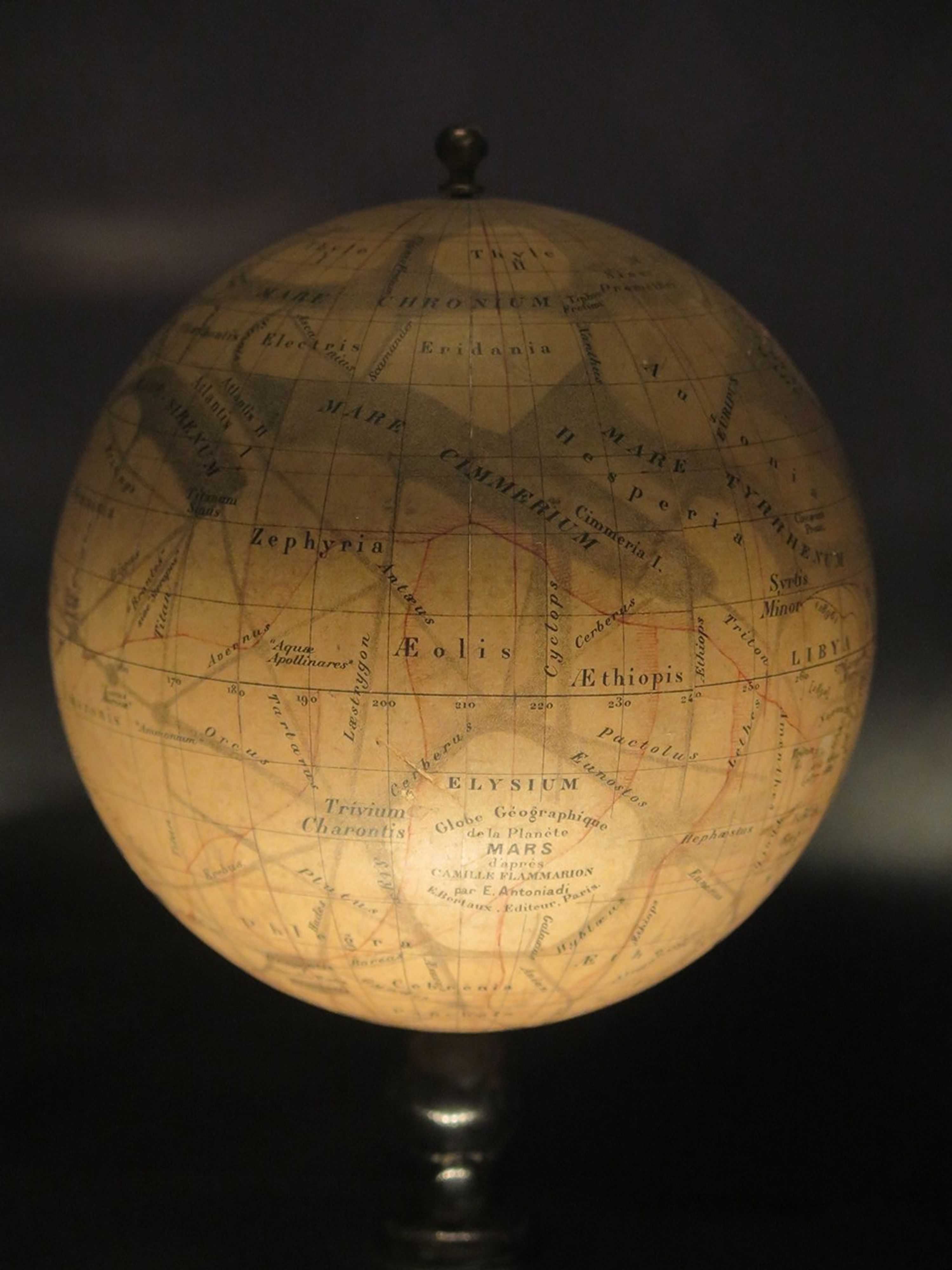 A color photo of a vintage globe of Mars, which appears golden yellow, and is crisscrossed with faint gray lines, and features wavy larger regions of gray near the top. All the markings are labeled with names in black type.