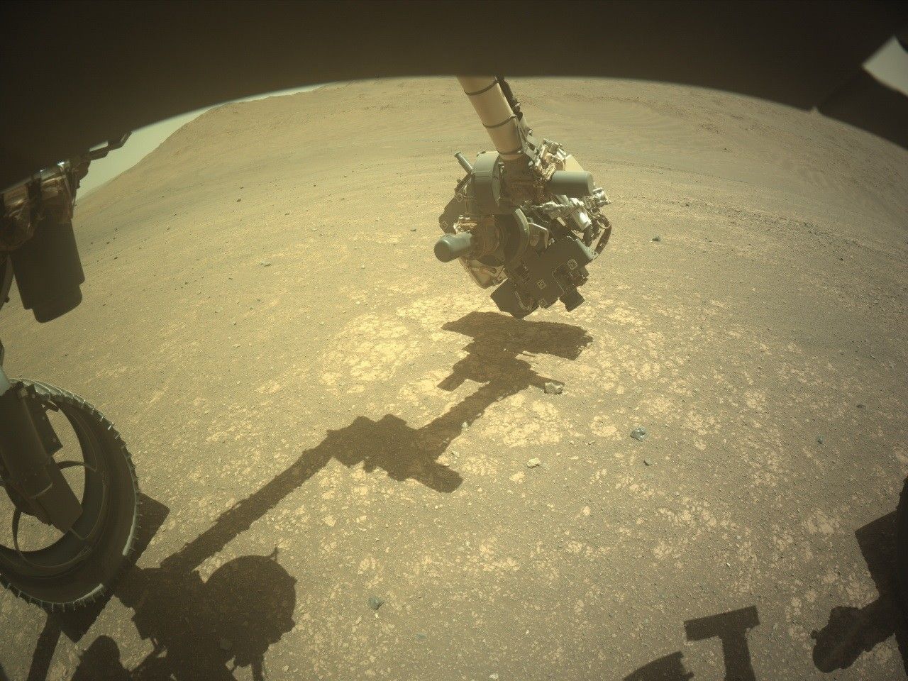 A color photo from the Mars surface shows pale orange, gritty terrain with lighter-colored flat rocks visible above the soil in several areas. At the image center, the Perseverance rover’s mechanical arm, with a boxy instrument at its end, extends from beyond the top of the frame and reaches toward the ground. Its shadow runs along the ground from the center of the frame to the lower left corner of the image. Shadowed portions of the rover are visible along the top of the image frame, and the lower-right corner, while a rover wheel is visible in the lower left.