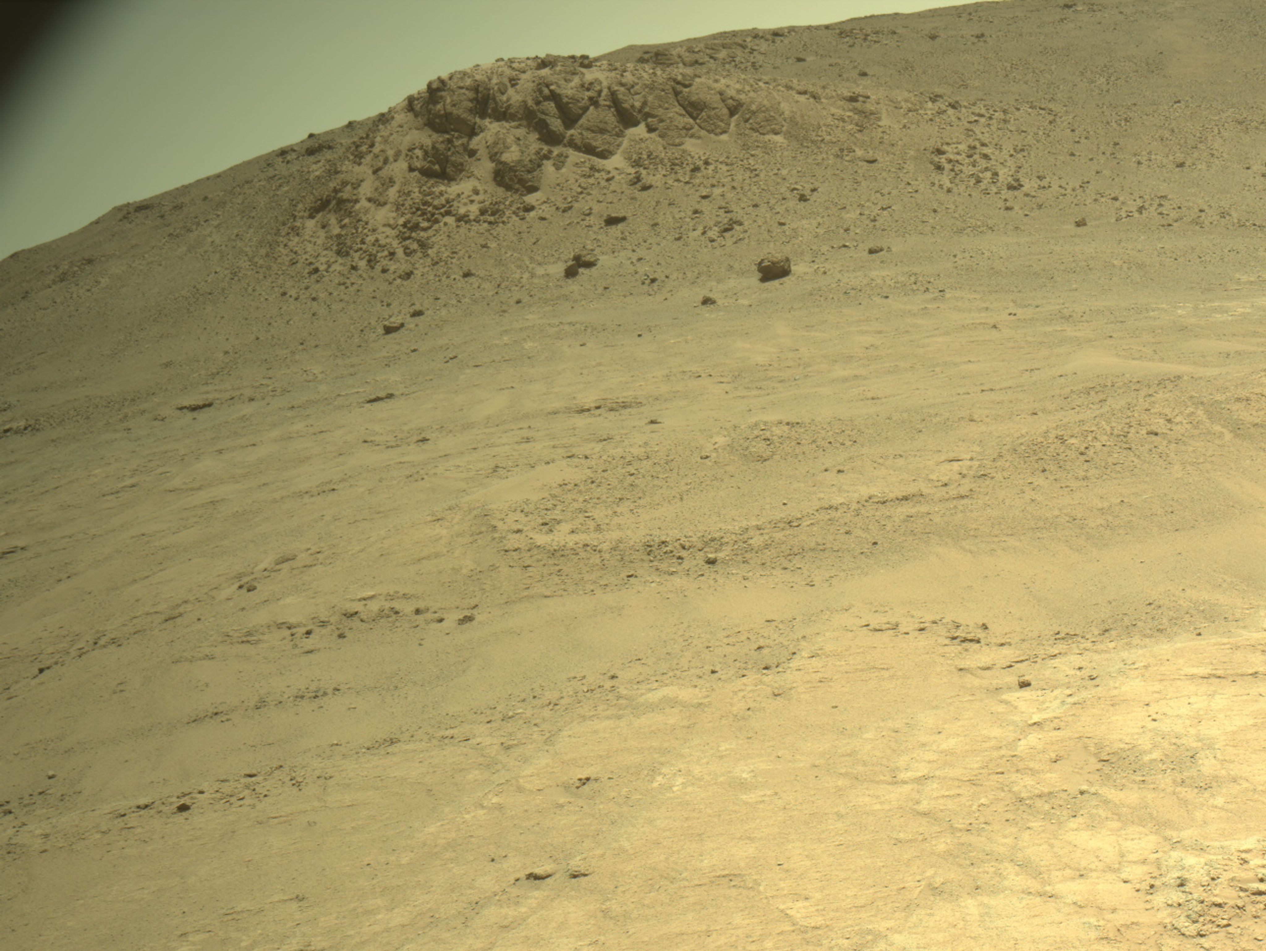 A color image from the Martian surface shows uneven yellow-tan ground leading up to the base of a hill, where the smoother ground dotted with small rocks turns into an outcropping of much larger rocks. That outcropping is on the horizon, which runs diagonally in the image, from the upper left side to the top right of the frame.