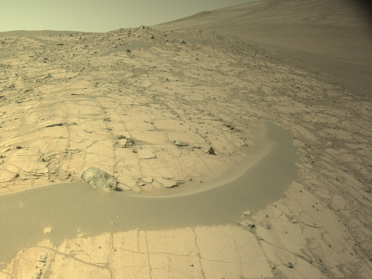 A color image from the Martian surface shows uneven yellow-tan ground that rises from the foreground upward toward the upper right corner of the image and off the frame. The ground appears dry, with narrow cracks dividing the ground into squares and other angular shapes. A narrow strip of smoother ground, like a low berm of mounded sand, curves from the left side toward the middle of the image. Beyond that, toward the upper left, the dry ground is more gravel-covered.