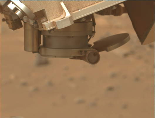 A close-up photo of an instrument on the Perseverance rover shows — against a blurred background of light tan-orange terrain — a metallic structure at the top of the frame, dusted with fine orange powder in some places. Pointing down from that is a short, barrel-shaped extension. Attached to the bottom of that is a “D”-shaped cover, attached to the mouth of the barrel on the right by a cylindrical hinge, fully opened toward the right, to an angle greater than 180 degrees.
