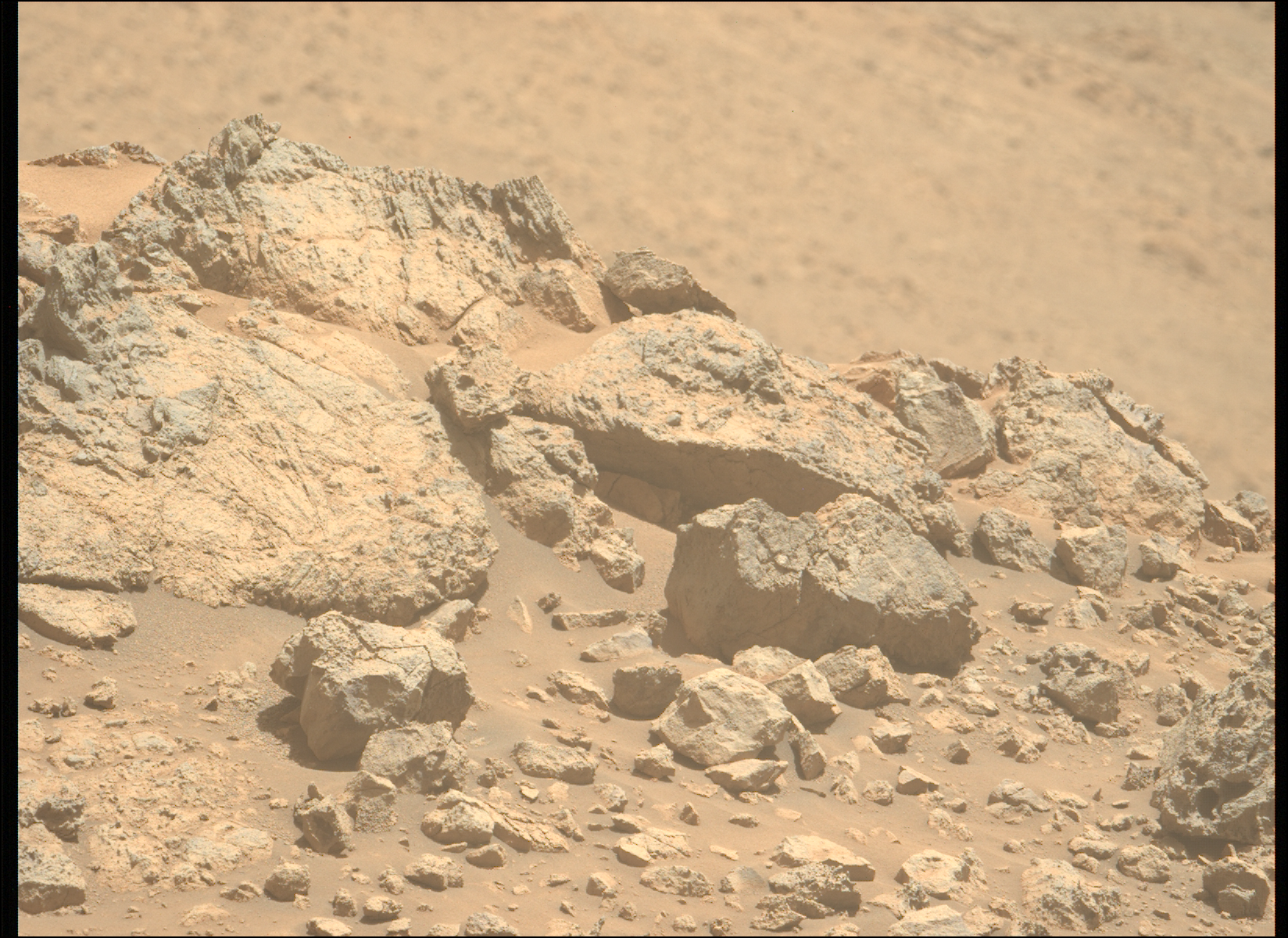 A color photo from the Martian surface shows pale orange, very rocky terrain in the foreground, taking up the lower two-thirds of the frame, creating a horizon line from the upper left corner of the image to the middle right side. The terrain beyond, in the distance, is blurry and takes up the rest of the frame. The rocks in the foreground range from tiny, sharp stones to large outcroppings, the latter showing some medium gray highlights among the orange rock and dust that comprise them. The largest rocks, on the left side, show numerous grooves and cracks on their faces, ranging from vertical to slightly left-skewing.