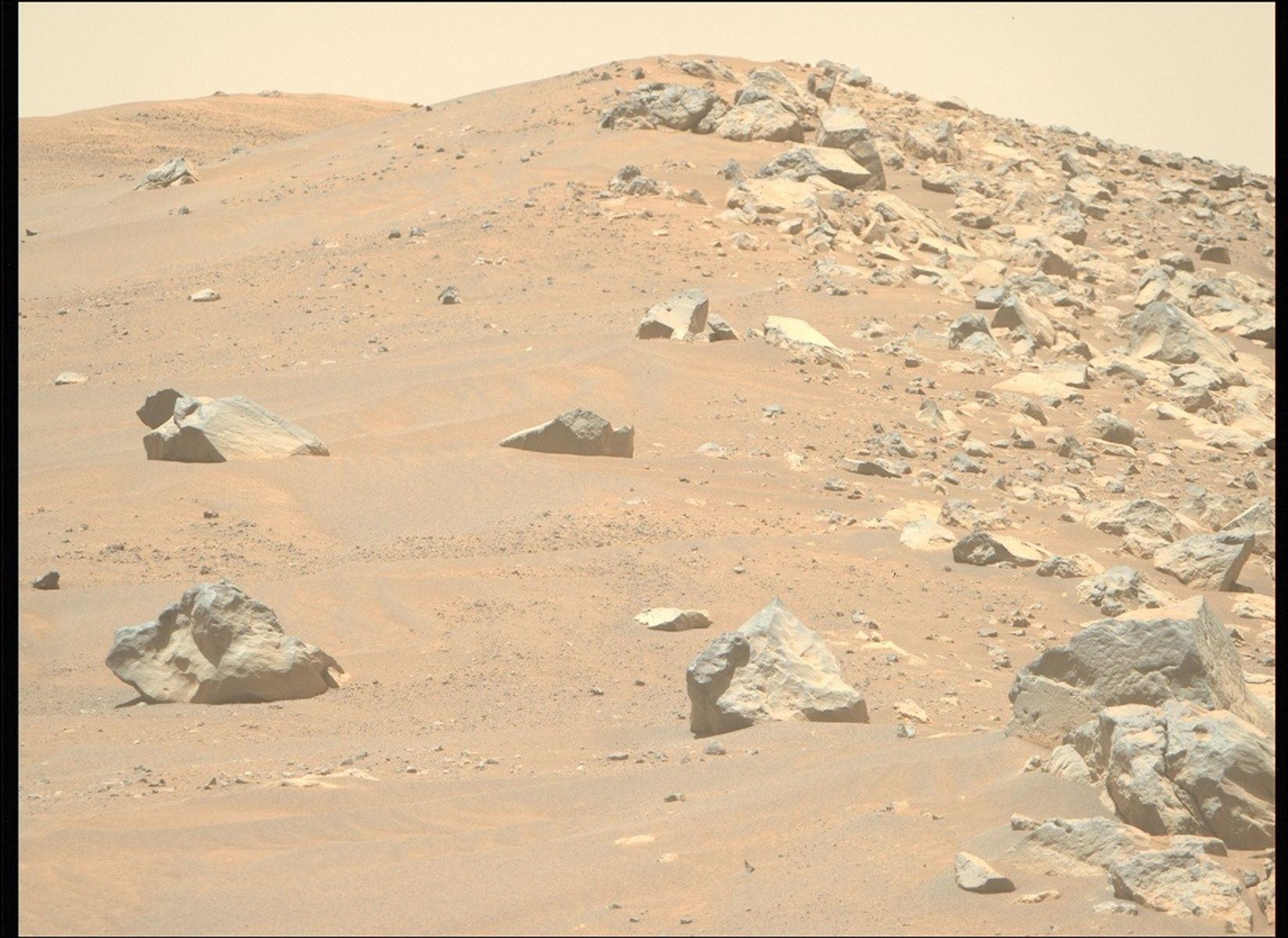 A color photo from the Martian surface shows a landscape of sandy, rocky terrain. What looks like a pair of light orange-brown sand dunes rise in the background. Four or five large, sharp-edged rocks are strewn among the sand on the left side of the image. But a clear vertical dividing line extends from the center of the image to the right side, where the sand is covered by similar rocks of all sizes. A pale yellow sky is visible above the scene.