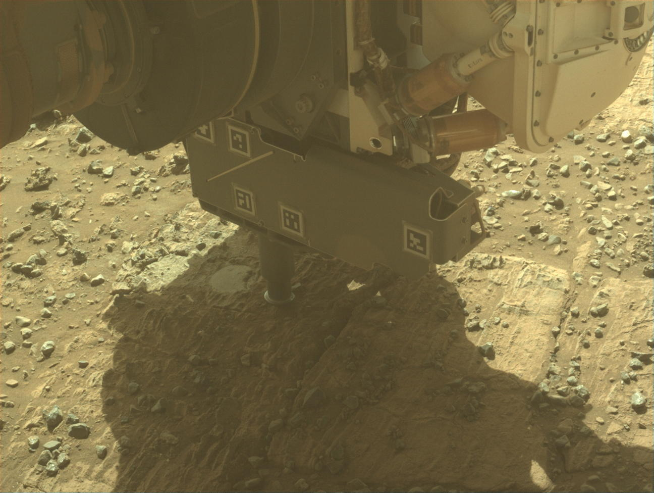 A color photo from the Mars surface shows pale yellow-orange, gritty terrain with lighter-colored, rectangular flat rocks visible above the soil. The turret at the end of the rover’s robotic arm dominates the upper half of the image. It shows two cylindrical probes reaching down to the ground below it.