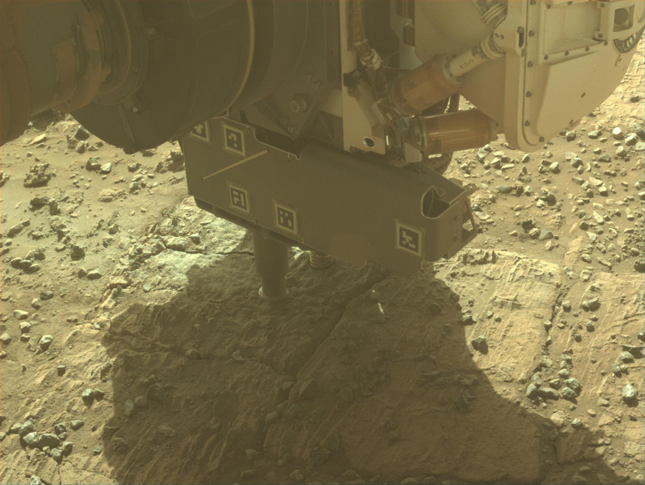 A color photo from the Mars surface shows pale yellow-orange, gritty terrain with lighter-colored, rectangular flat rocks visible above the soil. The turret at the end of the rover’s robotic arm dominates the upper half of the image. It shows two cylindrical probes reaching down to the ground below it.