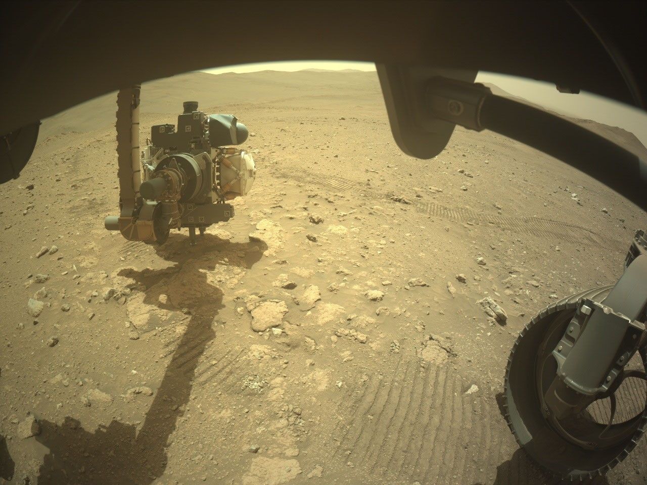 A wide-angle, slightly distorted view from beneath the Perseverance rover on Mars. Parts of the rover's dark chassis frame the top edge. On the bottom right, a section of one of its treaded wheels rests on the dirt. On the left, the rover's robotic arm, featuring a complex instrument turret at its end, is extended toward the ground. The arm casts a long, distinct shadow over the reddish-brown, rocky sand. Parallel rover tracks are pressed into the soil, leading away toward a relatively flat, barren landscape that ends at a horizon of distant, low hills under a pale sky.