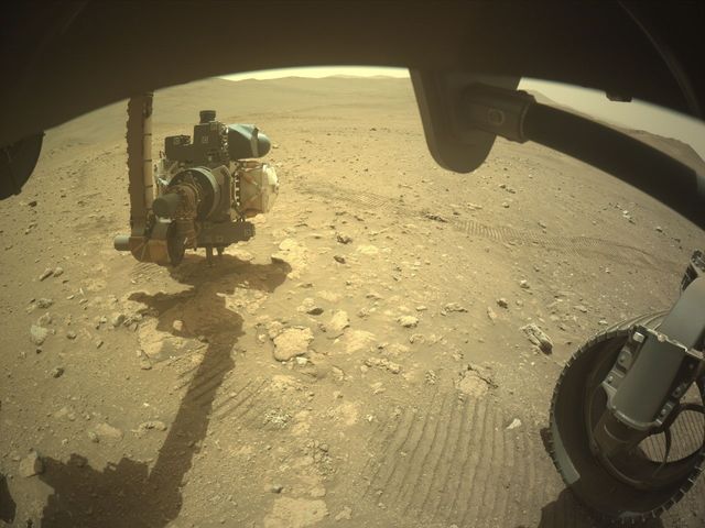 Perseverance strekt zijn instrumentenarm uit boven rotsen bij Idubi op Mars