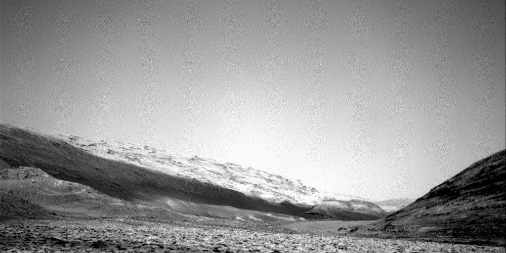 A grayscale landscape photo from Mars, showing a broad, medium gray, rock-strewn plain in the foreground extending far into the distance, surrounded by several hills. The closest hill, in the lower right corner, rises up toward the side of the frame, and is darker than the floor below. A little farther out, on the left, a gentle slope rises from the low middle of the frame to middle of the left side; it’s about the same color as the valley floor, except for a band at its farthest edge, which darkens abruptly as if covered in shadow. The terrain appears softer than the ground below, more sandy than rocky. Beyond that along the same rising plane is a much lighter area, with scattered tiny peaks that are darker – looking like the top of a baked meringue pie. The channel floor runs between these hills, then appears to curve toward the right, behind the first hill and in front of the second. Finally, one other rounded peak appears above the horizon, behind these other two hills in the lower right of the frame, all of this below a light-gray sky filling more than half of the frame above.