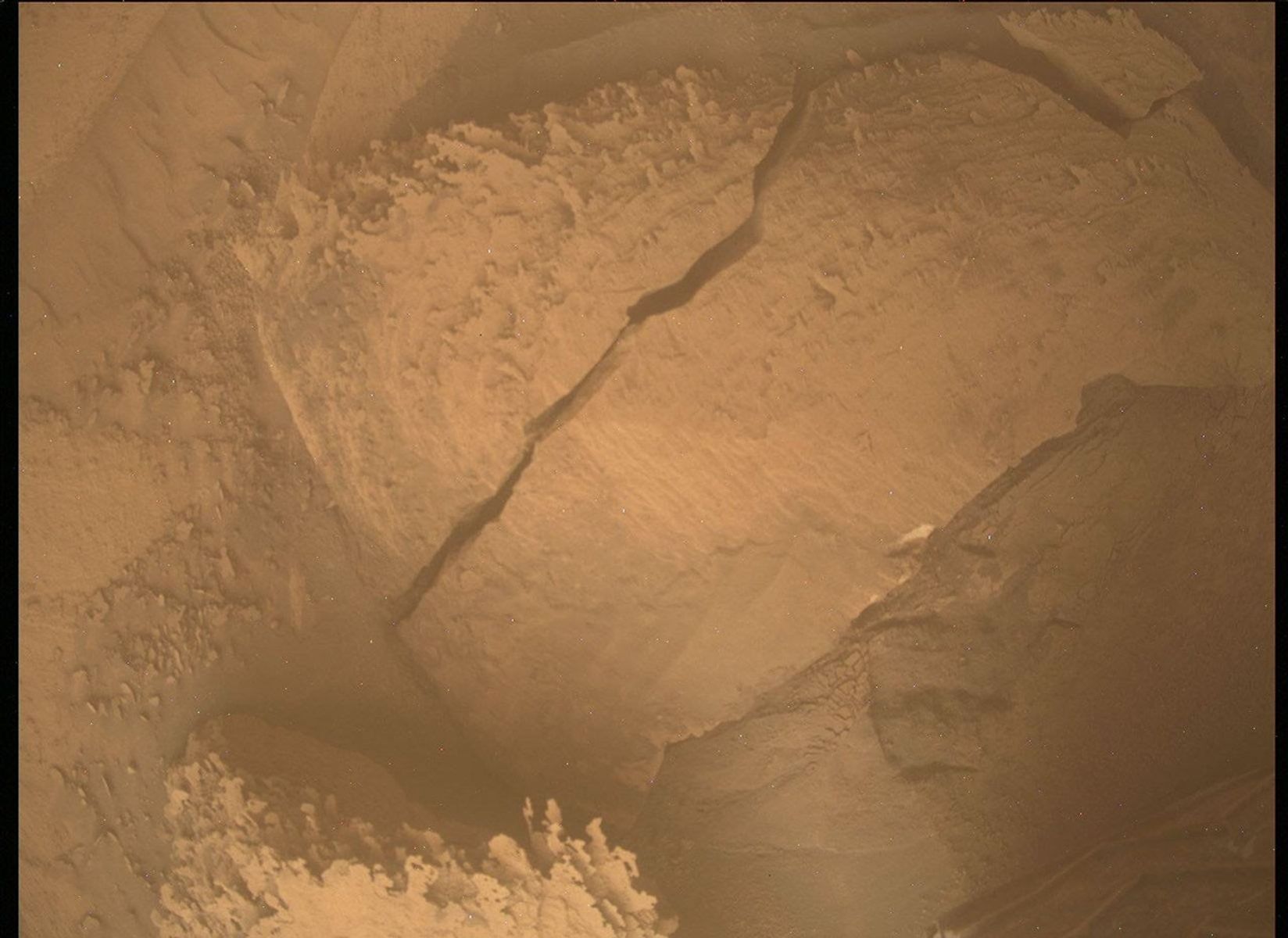 A color photograph from the Martian surface shows an overhead view of a small area of pale orange terrain, with an apparent slab of rock in the center of the image surrounded by fine, sandy soil and smaller rocks. At the center of the image is a long, wide crack in the slab, running from almost the top center of the frame toward the lower-left corner. Also, a small corner of the slab appears broken off, from an “L” shaped crack near the upper-right corner of the image.