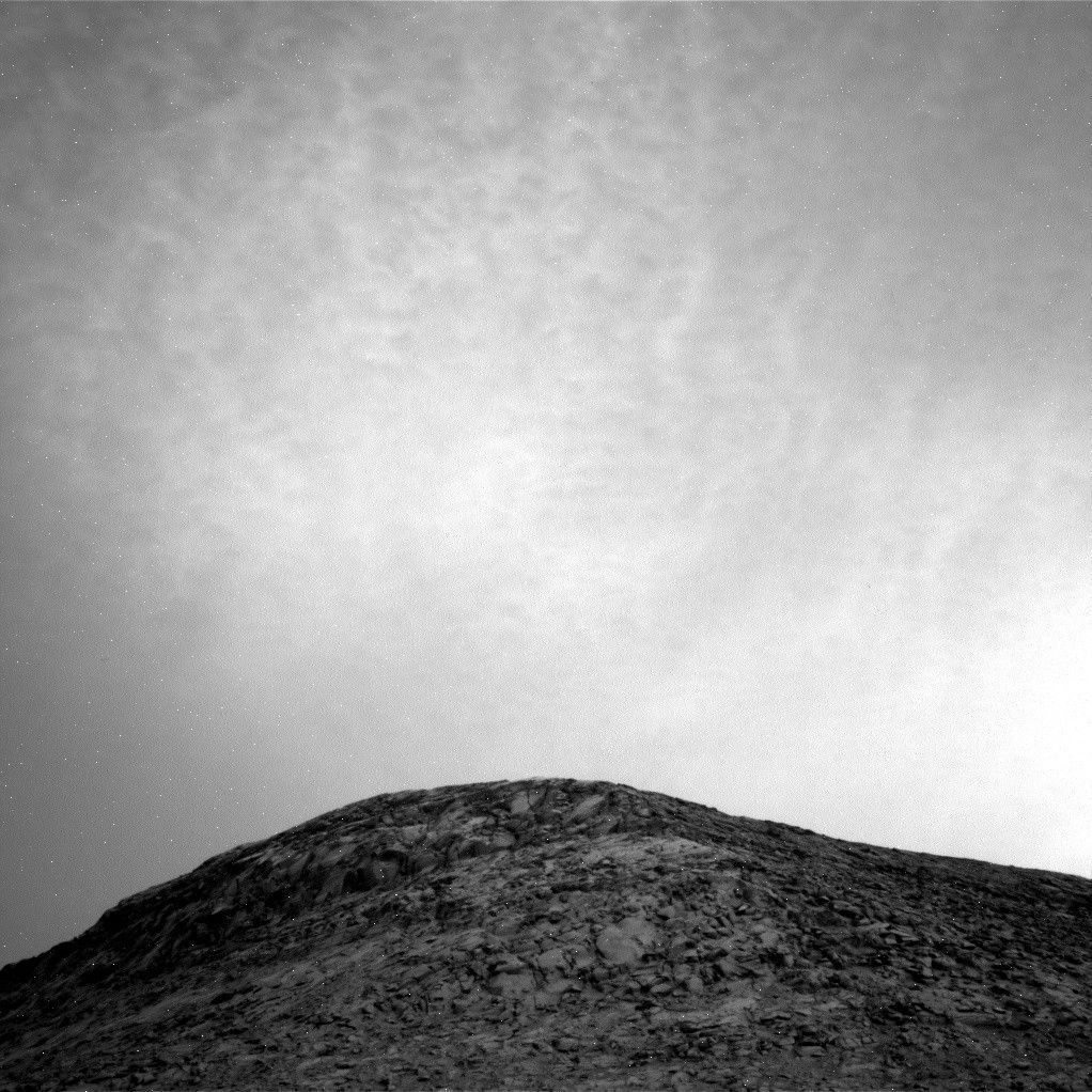 A grayscale image from the Martian surface shows a small gentle hill covered in dark gray rocky terrain, in the bottom quarter of the image. Above that is a light gray sky filled with glowing clouds resembling luminescent mother-of-pearl.