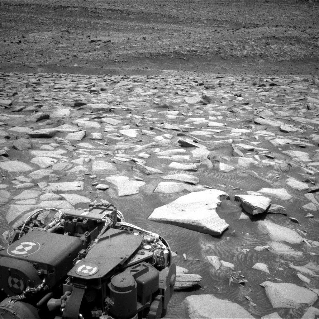 A grayscale photograph of the Martian surface shows a wide expanse of very rocky terrain, with sharp, angular stones arrayed as far as the eye can see, as if set together in a mosaic. The stones are very bright and light-toned, while the ground in between them is medium gray. A portion of the rover is visible in the foreground, in the lower left corner of the image.