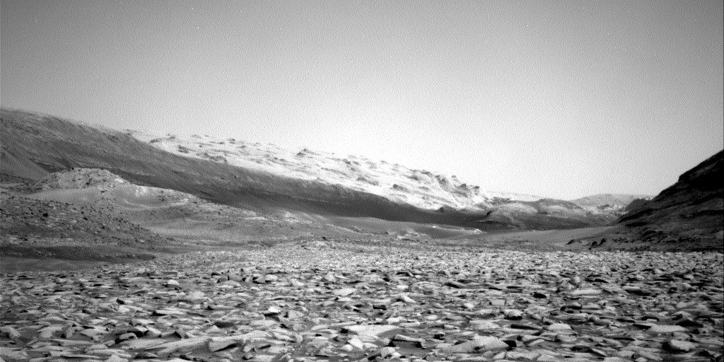 A grayscale photograph of the Martian surface shows a wide plain covered in sharp rocks, extending to the left and right edges of the frame, and far into the distance, all in shades of light gray. On either side of this channel, smoother hillsides rise at about 45 degree angles, and off on the horizon are an even higher range of hills. Those are much brighter gray, with dark areas, resembling the singed meringue peaks atop a lemon pie.
