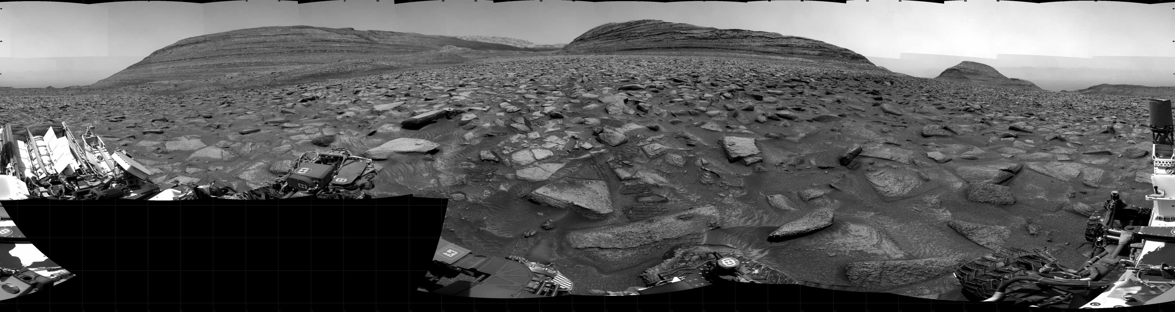 A grayscale panorama of the Martian surface shows a wide field of flat terrain dotted with flat, angular, medium-sized rocks, all in dark gray, stretching into the distance where a series of hills rise from the ground. A portion of the Curiosity rover is visible at the left and right sides of the image.