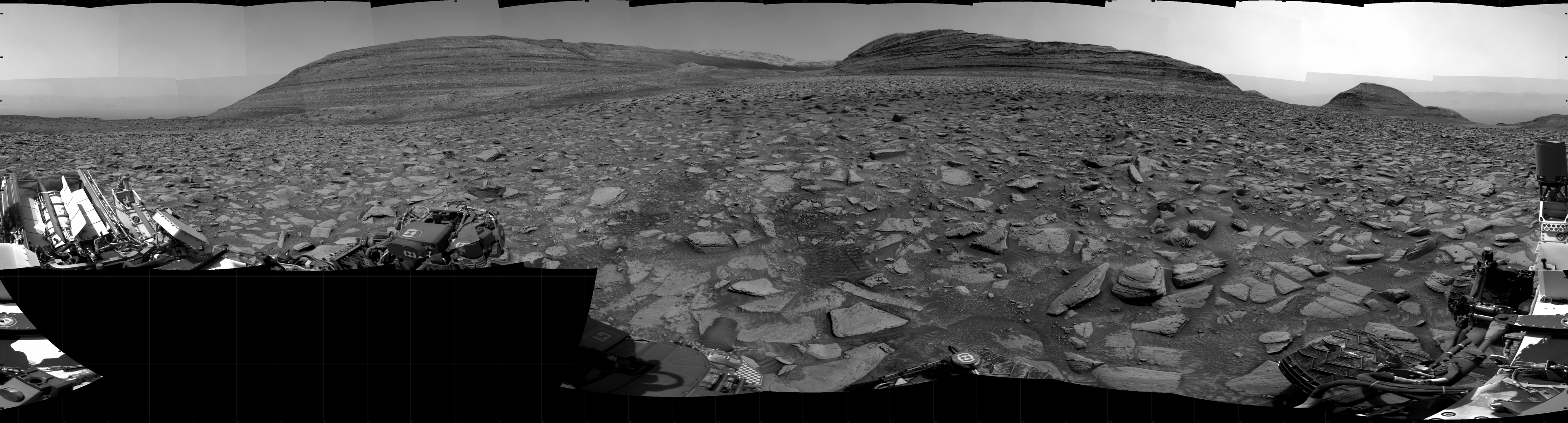 A grayscale panorama of the Martian surface shows a wide field of flat terrain dotted with flat, angular, medium-sized rocks, all in dark gray, stretching into the distance where a series of three hills rise from the ground, left to right on the horizon. A portion of the Curiosity rover is visible at the lower left corner of the image, and one wheel is visible in the lower right corner.