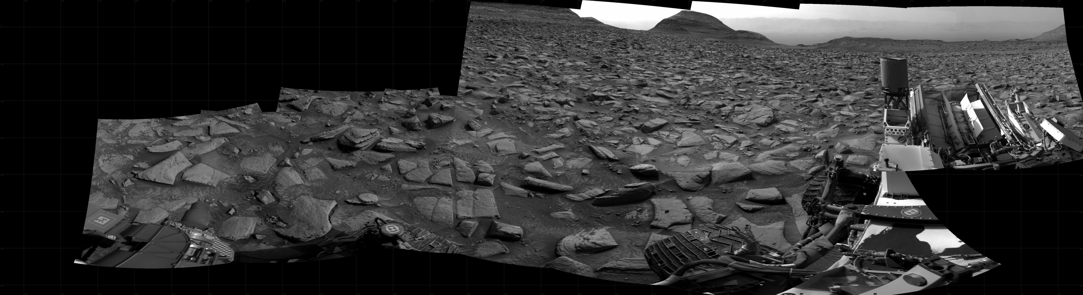 A grayscale panorama of the Martian surface shows a wide field of flat terrain dotted with flat, angular, medium-sized rocks, all in dark gray, stretching into the distance where a small hill rises from the ground. Features on the horizon are silhouetted by a bright, gray sky beyond, and what looks like a fog bank rests on the horizon beyond the hill, stretching horizontally about halfway up the visible sky. A portion of the top deck of the Curiosity rover and two of its wheels are visible at the right side and bottom of the frame; more of the rover is visible at the bottom left and lower left corner of the image.