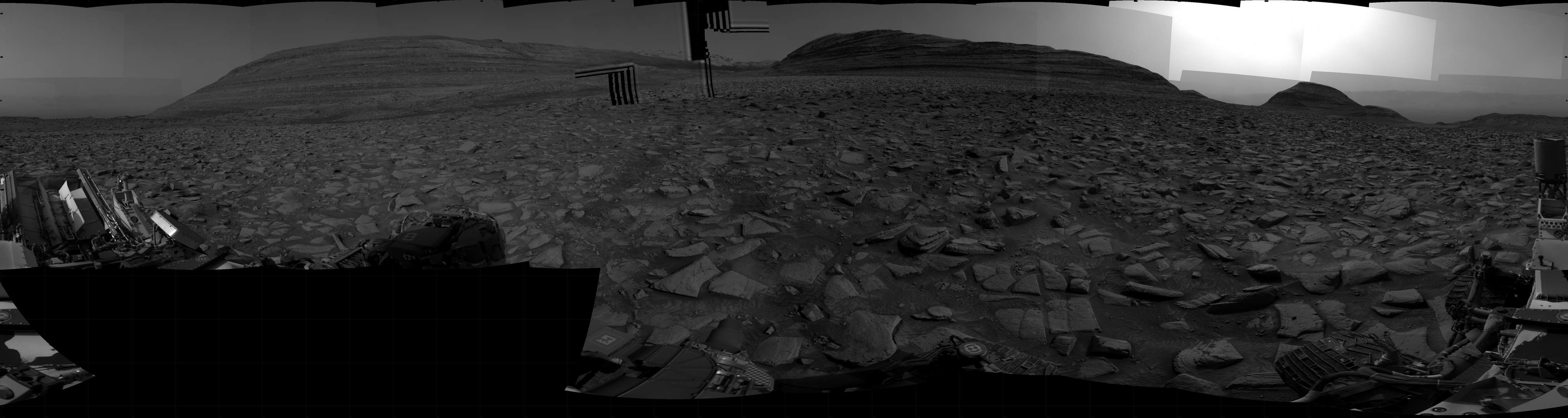 A grayscale panorama of the Martian surface shows a wide field of flat terrain dotted with flat, angular, medium-sized rocks, all in dark gray, stretching into the distance where three hills rise from the ground, with the ones at left and center being higher and wider than the one on the right. Features on the horizon are silhouetted by a gray sky beyond. A portion of the Curiosity rover and two of its wheels are visible at the bottom right corner of the frame; more of the rover is visible at the bottom and lower left corner of the image.