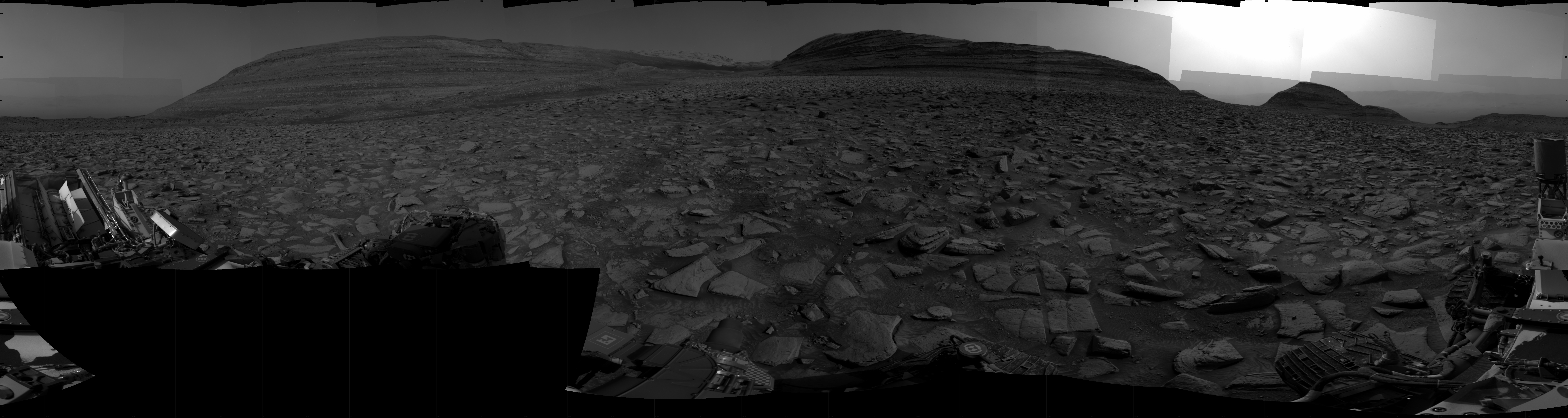 A grayscale panorama of the Martian surface shows a wide field of flat terrain dotted with flat, angular, medium-sized rocks, all in dark gray, stretching into the distance where three hills rise from the ground, with the ones at left and center being higher and wider than the one on the right. Features on the horizon are silhouetted by a gray sky beyond. A portion of the Curiosity rover and two of its wheels are visible at the bottom right corner of the frame; more of the rover is visible at the bottom and lower left corner of the image.