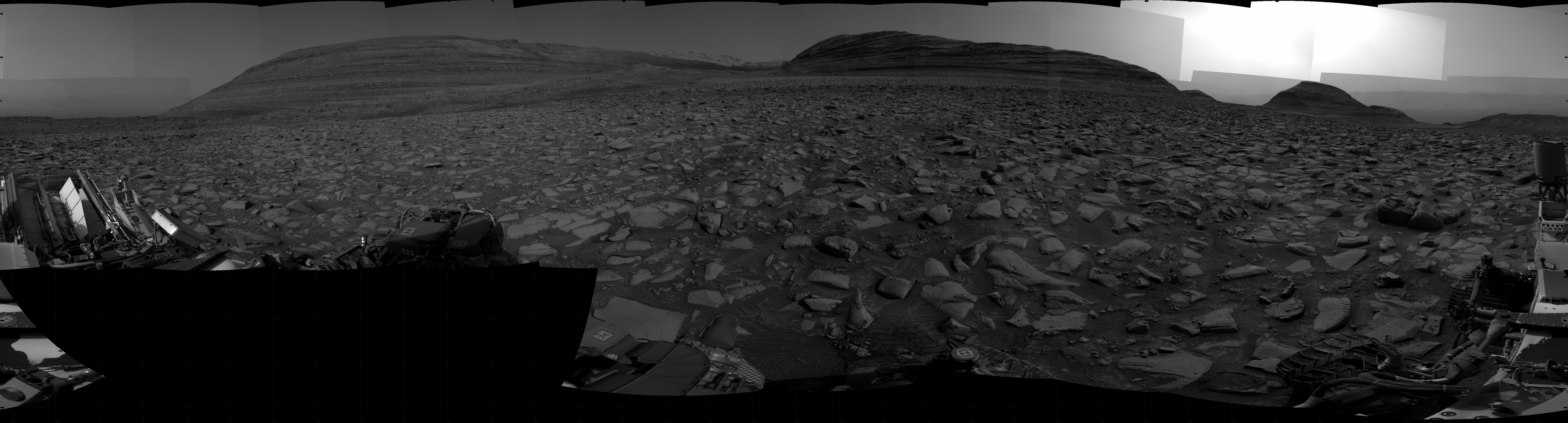A grayscale panorama of the Martian surface shows a wide field of flat terrain dotted with flat, angular, medium-sized rocks, all in dark gray, stretching into the distance where three hills rise from the ground, with the ones at left and center being higher and wider than the one on the right. Features on the horizon are silhouetted by a gray sky beyond. A portion of the Curiosity rover and two of its wheels are visible at the bottom right corner of the frame; more of the rover is visible at the bottom and lower left corner of the image.