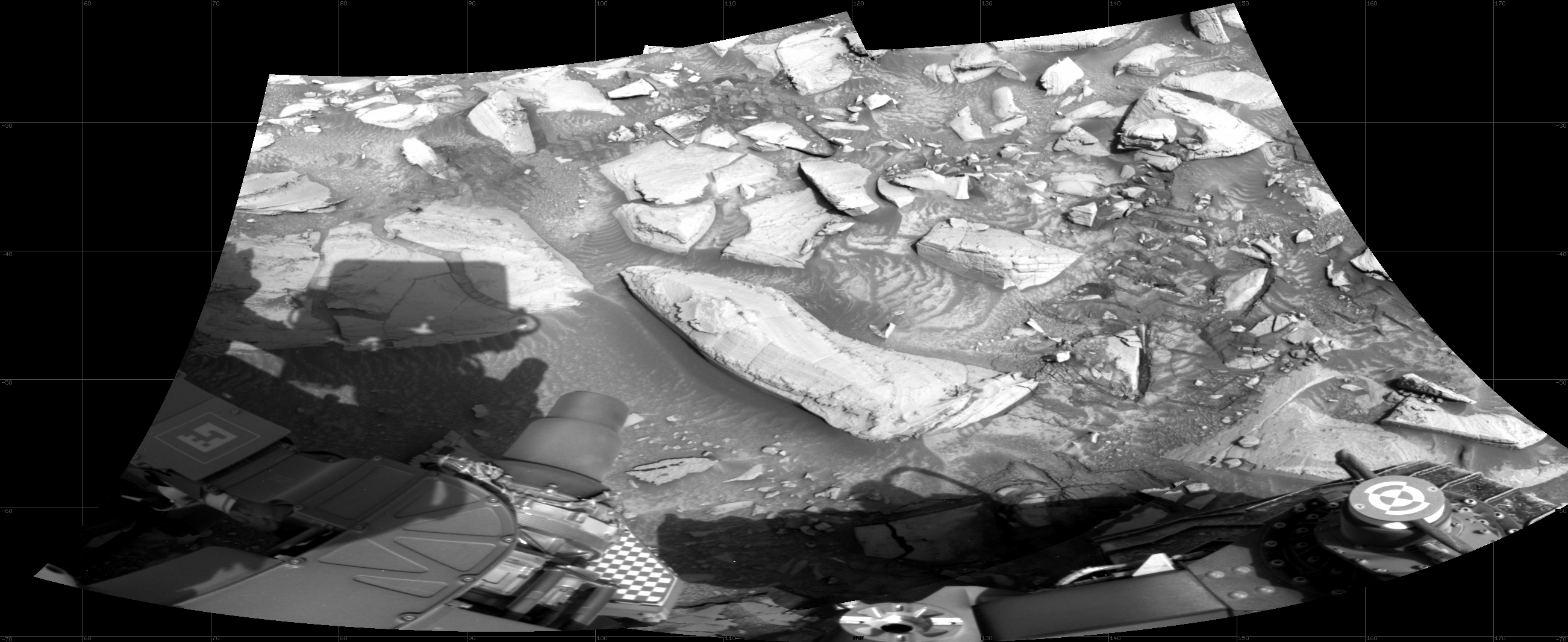 A grayscale image of the Curiosity rover workspace on Mars, showing medium-gray soil with numerous large, flat, lighter-toned rocks embedded in the ground. A portion of the rover is visible along the bottom of the image.