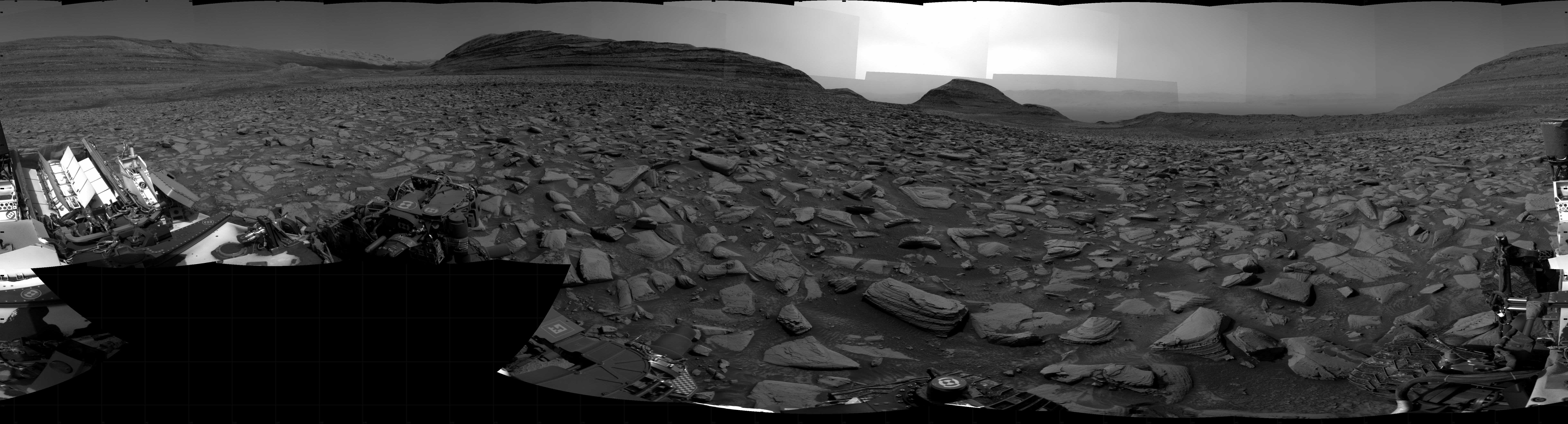 A grayscale panorama of the Martian surface shows a wide field of flat terrain dotted with flat, angular, medium-sized rocks, all in dark gray, stretching into the distance where a series of four hills rise from the ground, left to right on the horizon. Portions of the Curiosity rover are visible at the bottom of the image, including two wheels visible in the lower right corner and a third at the bottom center of the frame. The pale gray sky grows much brighter at center horizon, as if the Sun is either setting or rising.