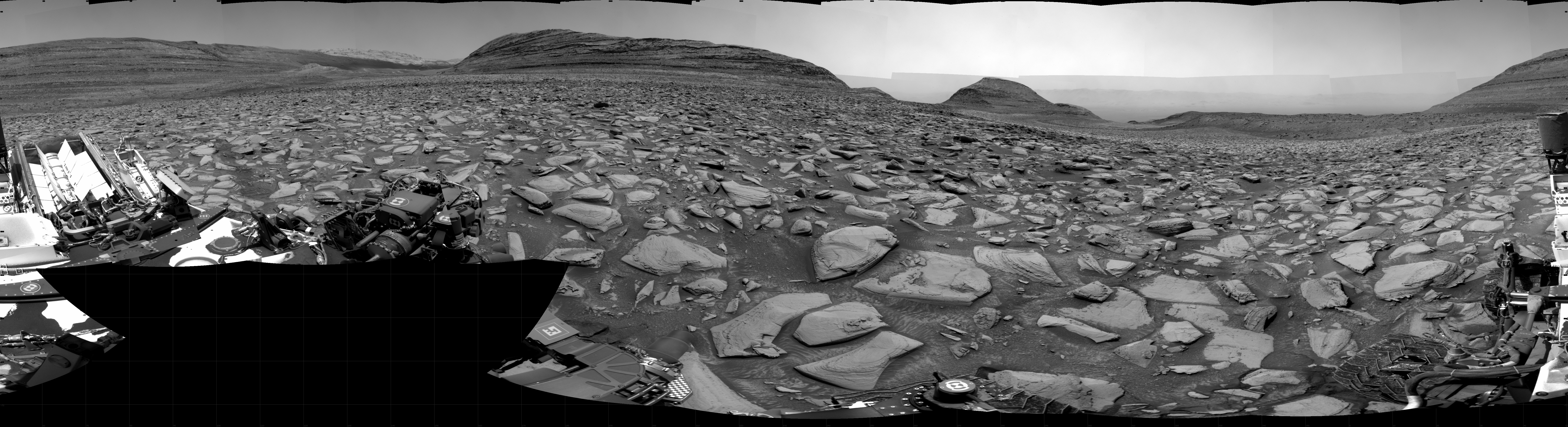 A grayscale panorama of the Martian surface shows a wide field of flat, dark gray terrain dotted with flat, angular, medium-sized rocks, all in lighter gray, stretching into the distance where a series of four hills rise from the ground, left to right on the horizon. Portions of the Curiosity rover are visible at the bottom of the image, including two wheels visible in the lower right corner and a third at the bottom center of the frame.