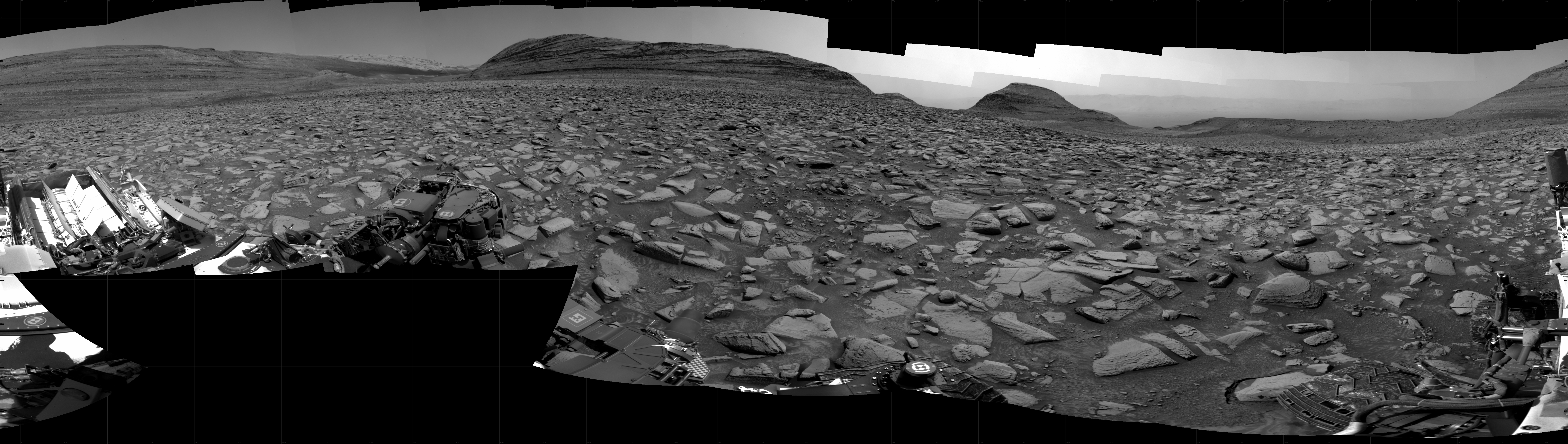 A grayscale panorama of the Martian surface shows a wide field of flat, dark gray terrain dotted with flat, angular, medium-sized rocks, all in lighter gray, stretching into the distance where a series of four hills rise from the ground, left to right on the horizon. Portions of the Curiosity rover are visible at the bottom of the image, including two wheels visible in the lower right corner and a third at the bottom center of the frame.