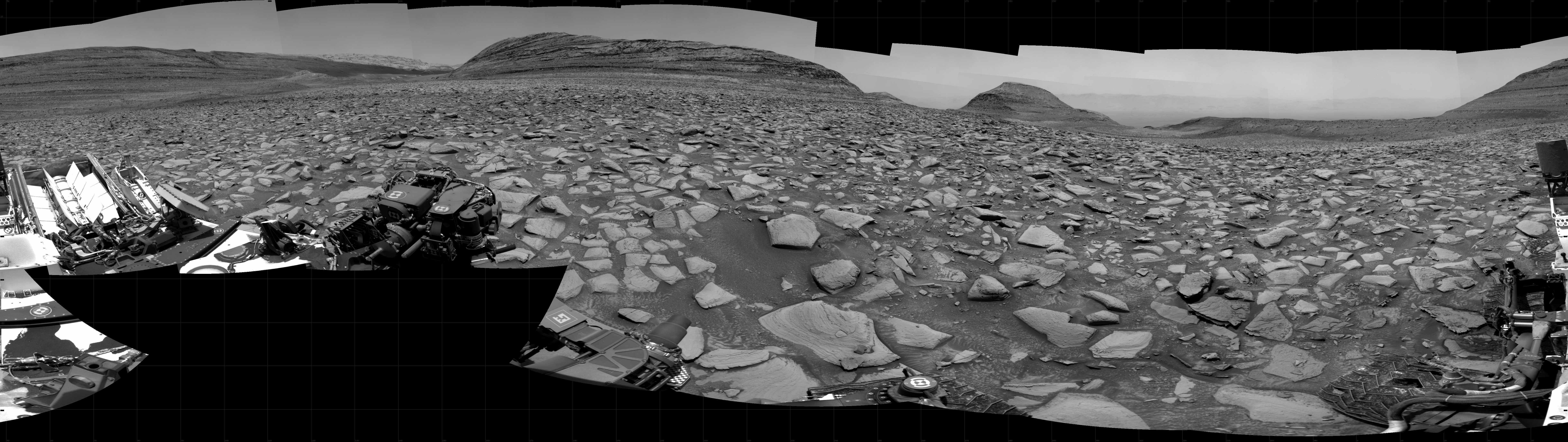 A grayscale panorama of the Martian surface shows a wide field of flat, dark gray terrain dotted with flat, angular, medium-sized rocks, all in lighter gray, stretching into the distance where a series of four hills rise from the ground, left to right on the horizon. Portions of the Curiosity rover are visible at the bottom of the image, including two wheels visible in the lower right corner and a third at the bottom center of the frame.
