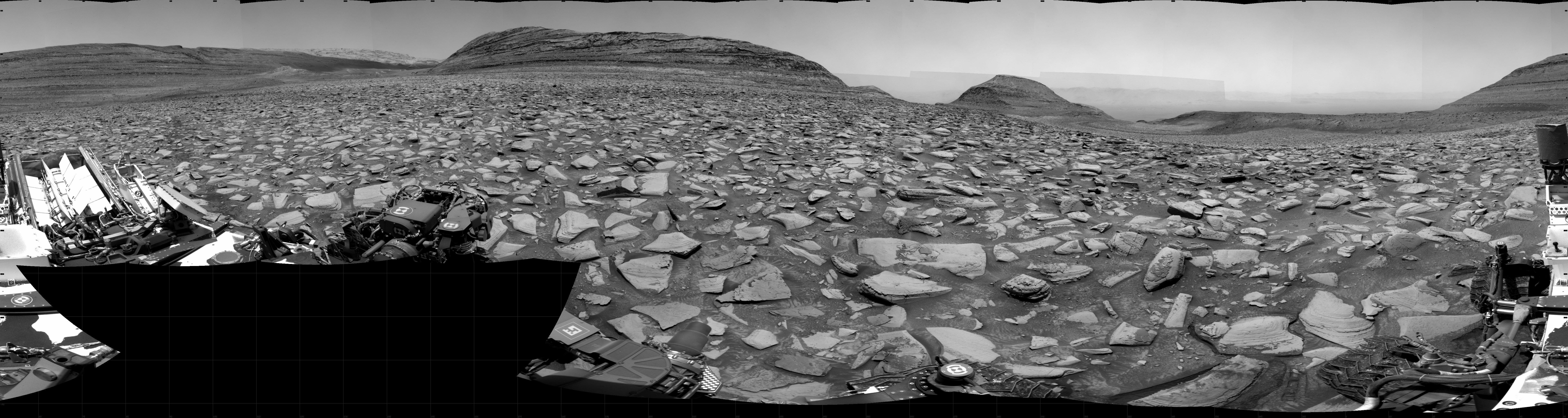 A grayscale panorama of the Martian surface shows a wide field of flat, dark gray terrain dotted with flat, angular, medium-sized rocks, all in lighter gray, stretching into the distance where a series of four hills rise from the ground, left to right on the horizon. Portions of the Curiosity rover are visible at the bottom of the image, including two wheels visible in the lower right corner and a third at the bottom center of the frame.