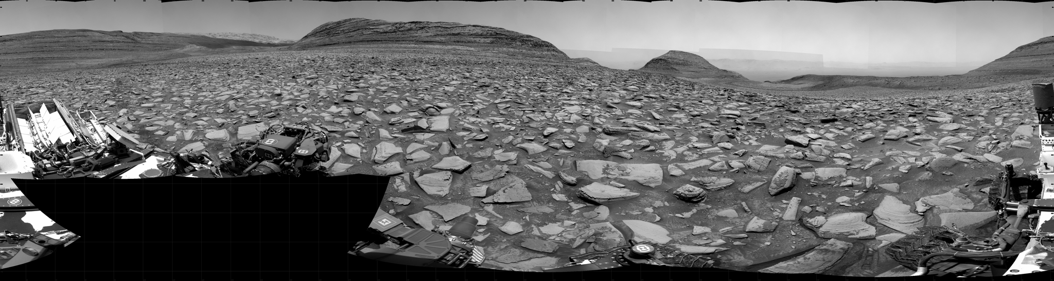 A grayscale panorama of the Martian surface shows a wide field of flat, dark gray terrain dotted with flat, angular, medium-sized rocks, all in lighter gray, stretching into the distance where a series of four hills rise from the ground, left to right on the horizon. Portions of the Curiosity rover are visible at the bottom of the image, including two wheels visible in the lower right corner and a third at the bottom center of the frame.