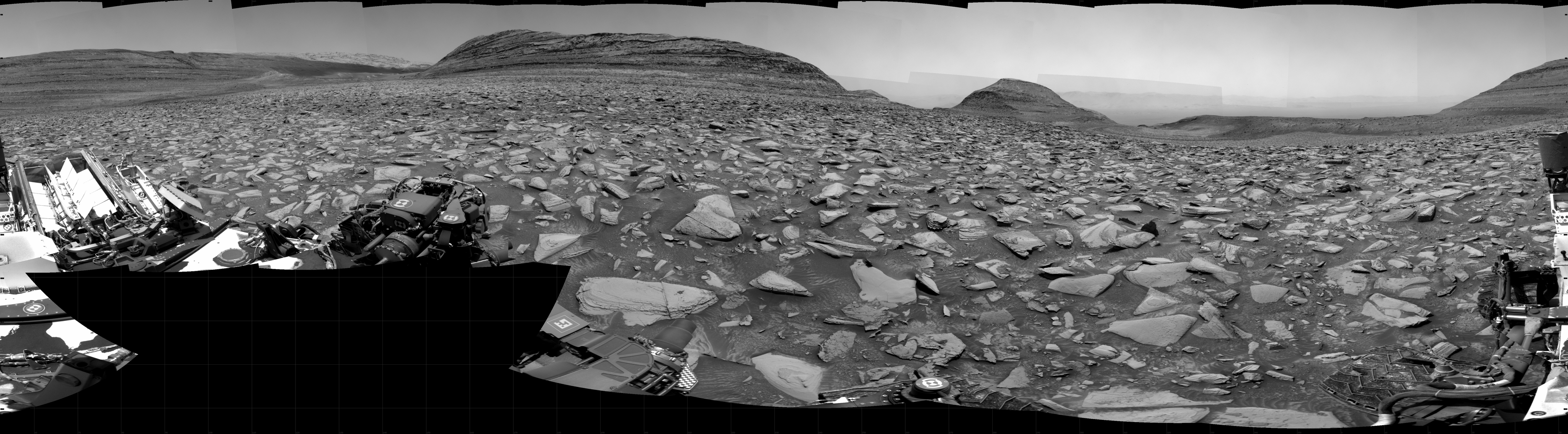 A grayscale panorama of the Martian surface shows a wide field of flat, dark gray terrain dotted with flat, angular, medium-sized rocks, all in lighter gray, stretching into the distance where a series of four hills rise from the ground, left to right on the horizon. Portions of the Curiosity rover are visible at the bottom of the image, including two wheels visible in the lower right corner and a third at the bottom center of the frame.