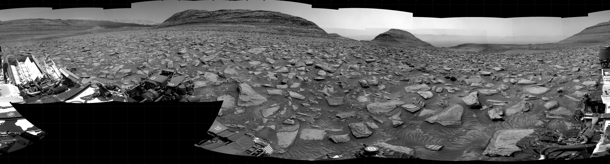 A grayscale panorama of the Martian surface shows a wide field of flat, dark gray terrain dotted with flat, angular, medium-sized rocks, all in lighter gray, stretching into the distance where a series of four hills rise from the ground, left to right on the horizon. Portions of the Curiosity rover are visible at the bottom of the image, including two wheels visible in the lower right corner and a third at the bottom center of the frame.