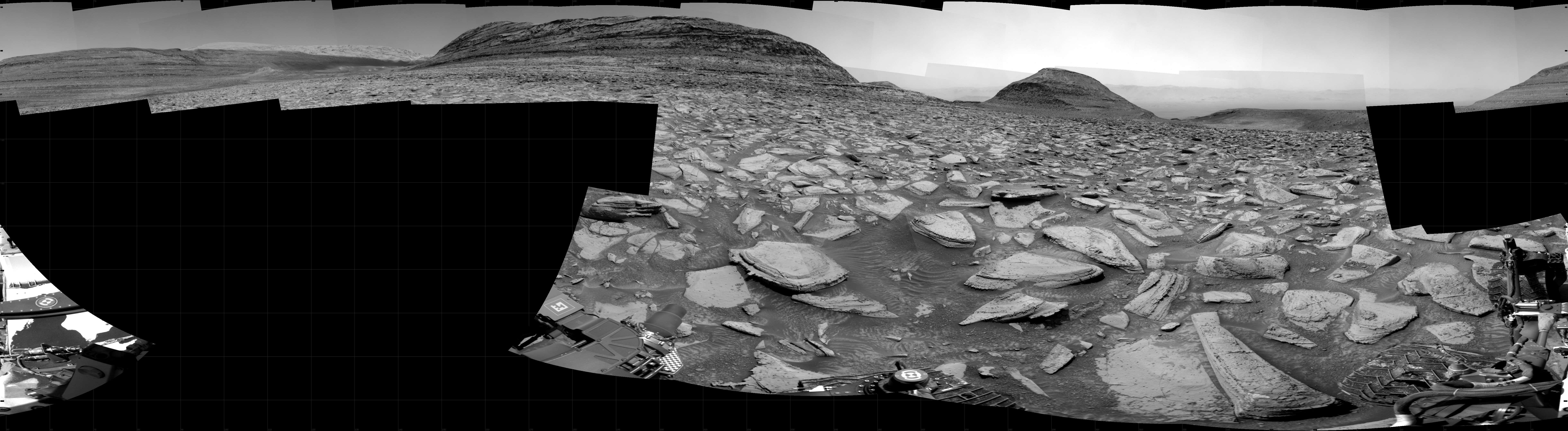 A grayscale partial panorama of the Martian surface shows a wide field of flat, dark gray terrain dotted with flat, angular, medium-sized rocks, all in lighter gray, stretching into the distance where a series of three hills rise from the ground, left to right on the horizon. Portions of the Curiosity rover are visible at the bottom of the image, including two wheels visible in the lower right corner of the frame.