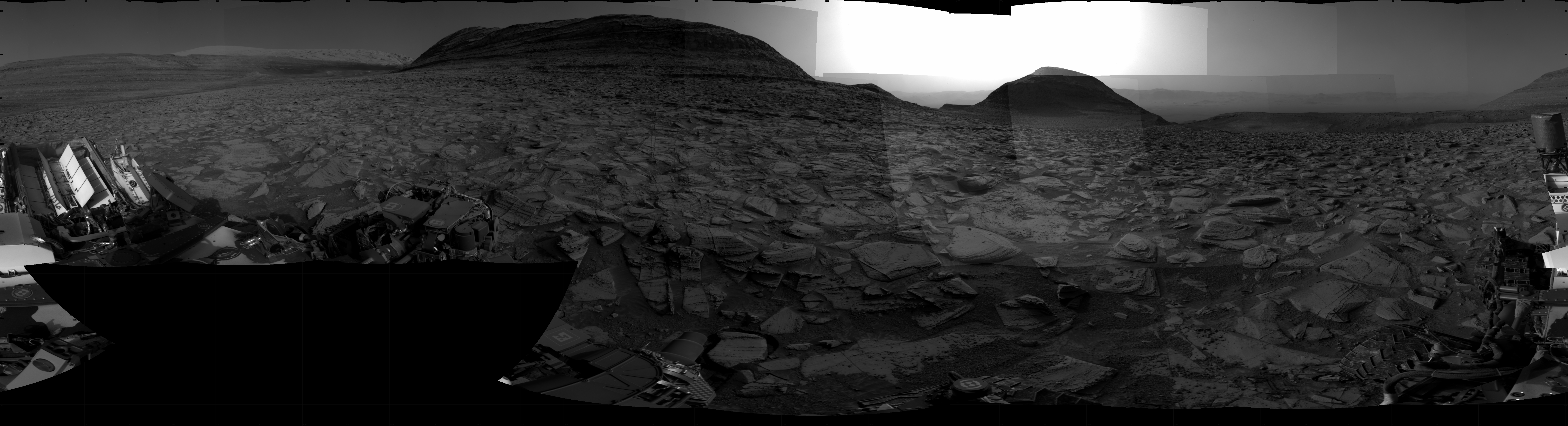 A grayscale panorama of the Martian surface shows a wide field of flat terrain dotted with flat, angular, medium-sized rocks, all in dark gray, stretching into the distance where a series of three hills rise from the ground, left to right on the horizon. Portions of the Curiosity rover are visible at the bottom of the image, including two wheels visible in the lower right corner of the frame, and a third at the bottom center of the frame. The pale gray sky grows much brighter at center horizon, as if the Sun is either setting or rising.