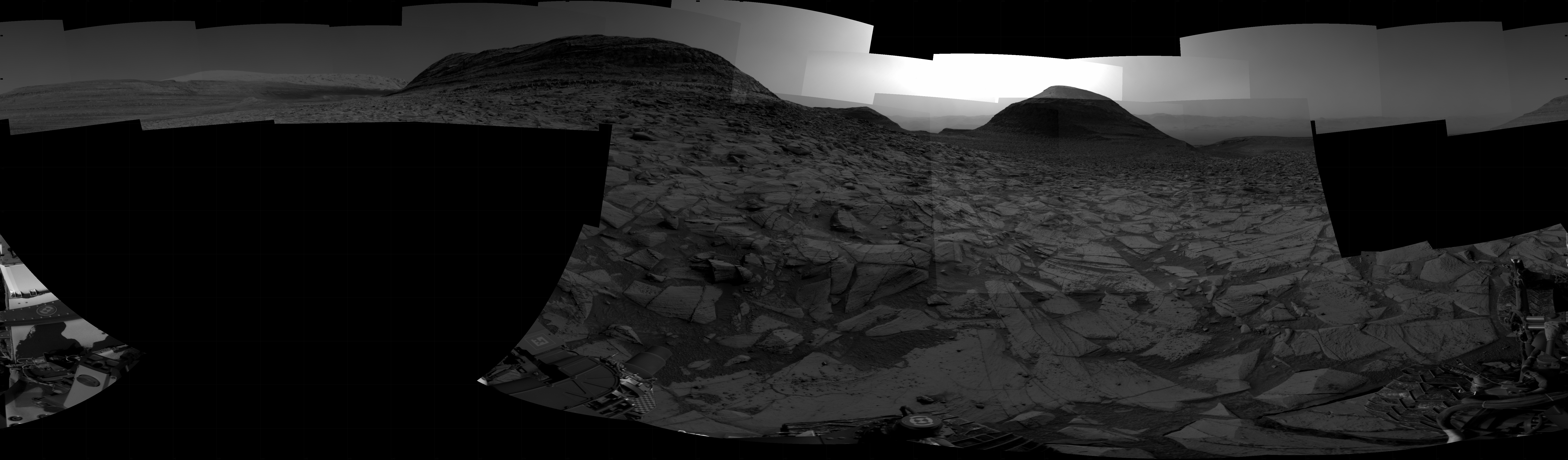 A grayscale panorama of the Martian surface, possibly at night or in twilight, shows a wide field of flat, dark gray terrain dotted with flat, angular, medium-sized rocks, also dark gray, stretching into the distance where a pair of hills rise from the ground on the horizon. A bright light illuminates a small part of the sky, between the two hills at the top center of the image. Portions of the Curiosity rover are visible at the bottom of the image, including a wheel visible in the lower right corner.