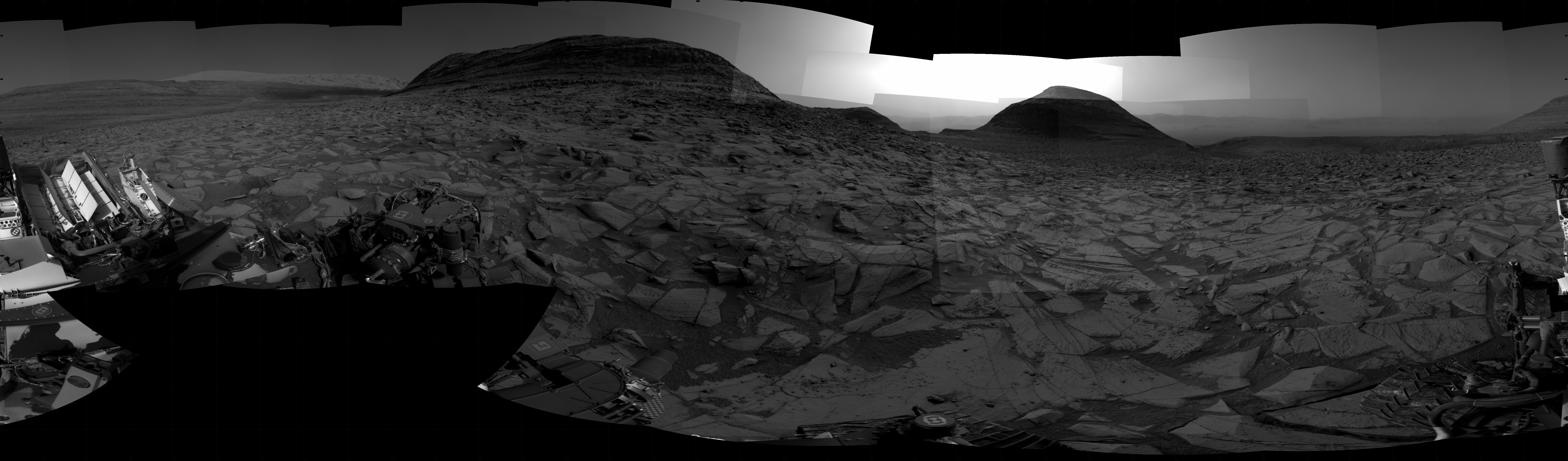 A grayscale panorama of the Martian surface, possibly at night or in twilight, shows a wide field of flat, dark gray terrain dotted with flat, angular, medium-sized rocks, also dark gray, stretching into the distance where a pair of hills rise from the ground on the horizon. A bright light illuminates a small part of the sky, between the two hills at the top center of the image. Portions of the Curiosity rover are visible at the bottom of the image, including a wheel visible in the lower right corner.