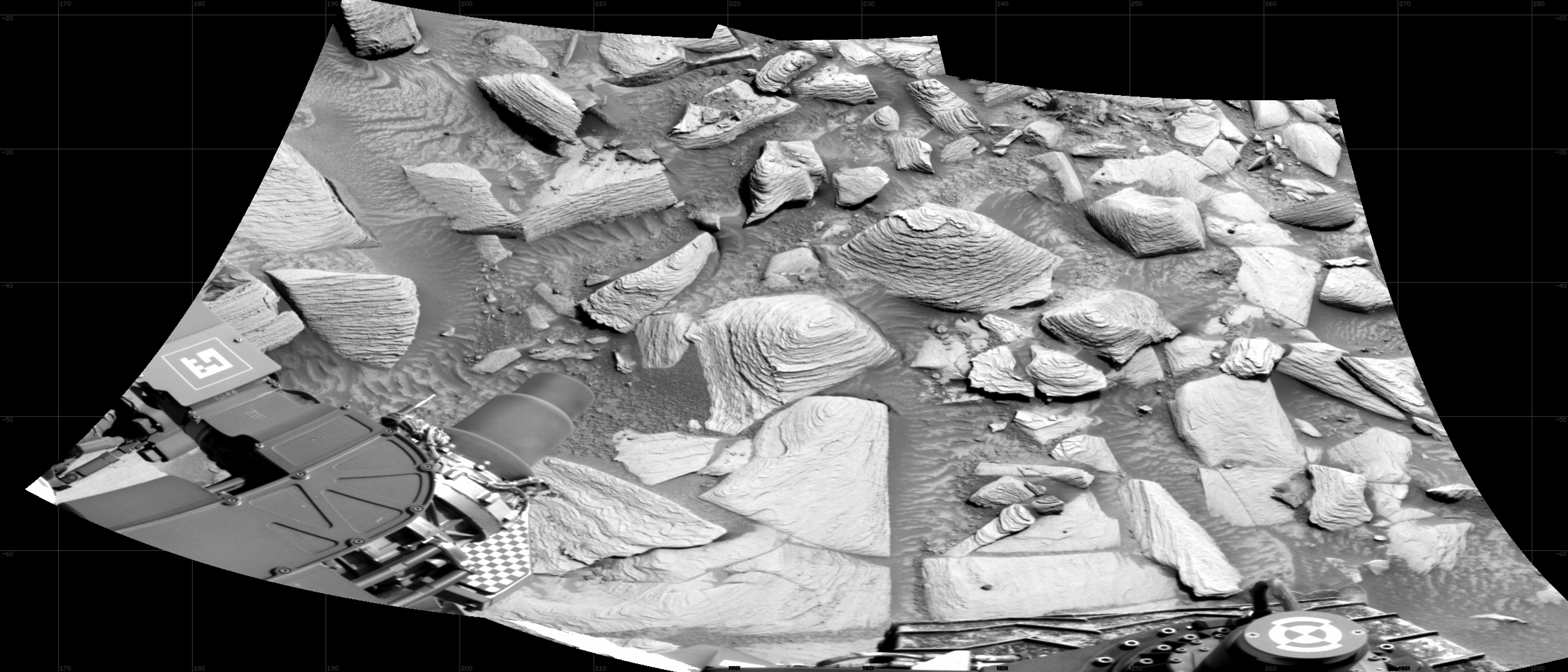 A grayscale, trapezoid-shaped image of the Martian surface shows a patch of very rocky terrain next to the Curiosity rover. Parts of the rover are visible at the bottom of the frame. Several of the large rocks pushing up from the ground are layered and pyramid shaped.