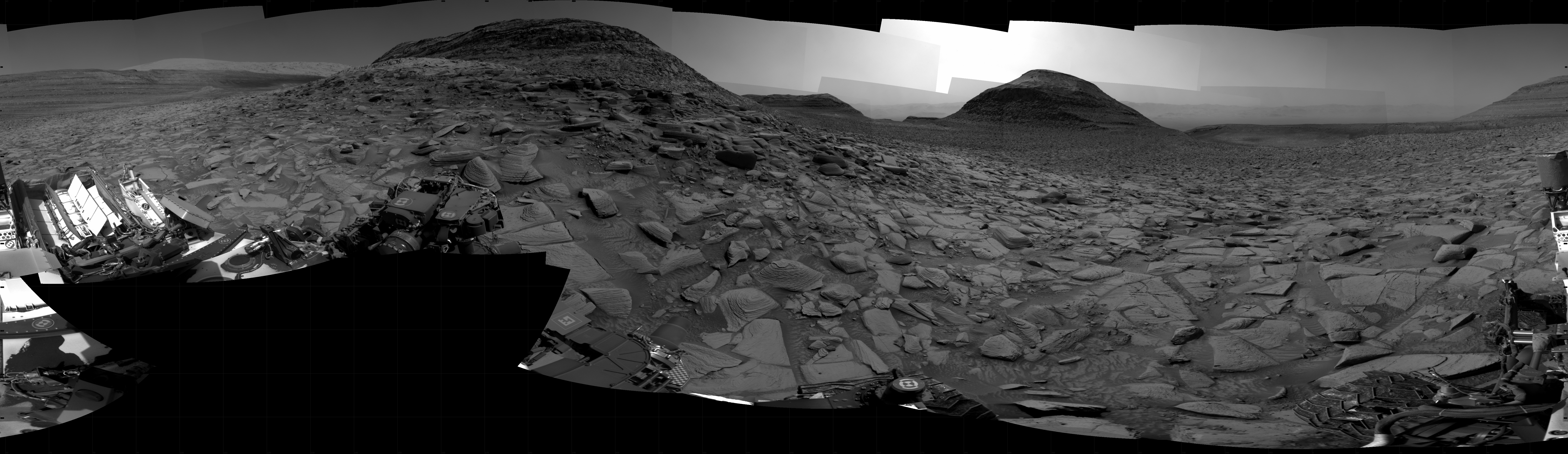 A grayscale panorama of the Martian surface, possibly at night or in twilight, shows a wide field of flat, dark gray terrain dotted with flat, angular, medium-sized rocks, lighter gray, stretching into the distance where a pair of hills rise from the ground on the horizon. A bright light illuminates a small part of the sky, off the frame and above the smaller hill on the right. A third hill begins to slope upward on the horizon, at the right edge of the frame. Portions of the Curiosity rover are visible at the bottom of the image, including a wheel visible in the bottom center of the frame, and two others in the lower right corner.