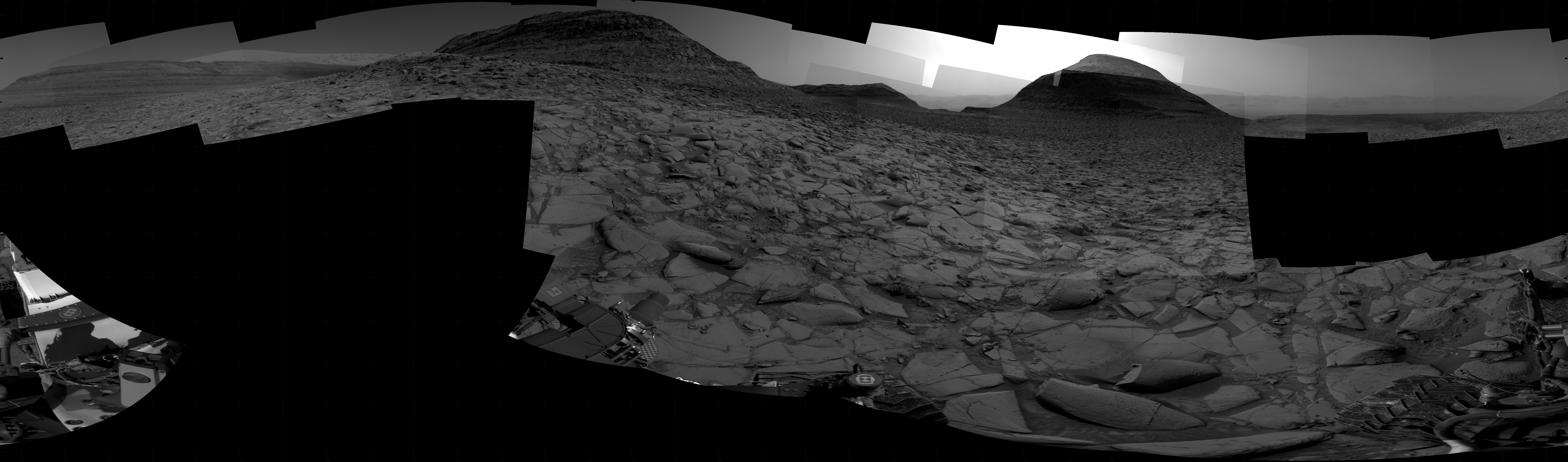 A grayscale panorama of the Martian surface shows a wide field of flat, dark gray terrain dotted with flat, angular, medium-sized rocks stretching into the distance where a series of three hills rise from the ground, left to right on the horizon, the middle one lower on the horizon and farther away than the others. Portions of the Curiosity rover are visible at the bottom of the image, including one wheel visible in the lower right corner and another at the bottom center of the frame.